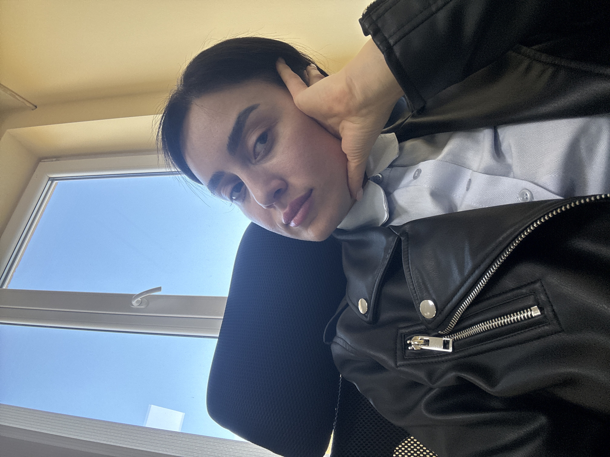 A person is sitting in a chair by a window. The person is wearing a light blue collared shirt and a black leather jacket. Their left hand is touching their face. The window is showing a blue sky. The location is likely inside a building in Tbilisi, Georgia, based on the architectural style of the window and the person's attire, consistent with urban environments in the region.
