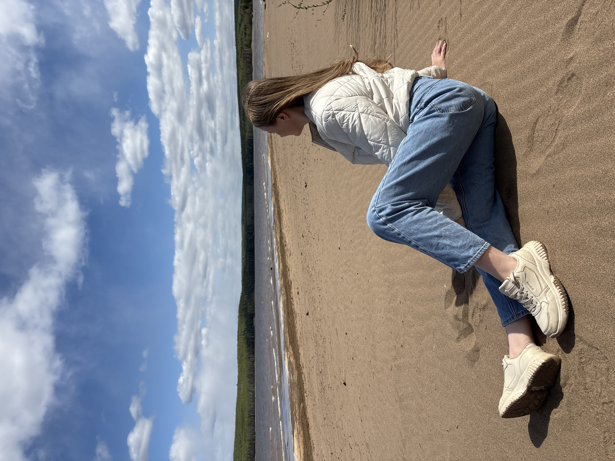 A young woman is lying on a sandy beach. She is wearing a white quilted jacket, blue jeans, and off-white sneakers. She appears to be looking toward the water. The beach borders a body of water with a forest on the opposite shore. The sky is blue with white clouds. The location is likely in Chaikovsky, Russia.