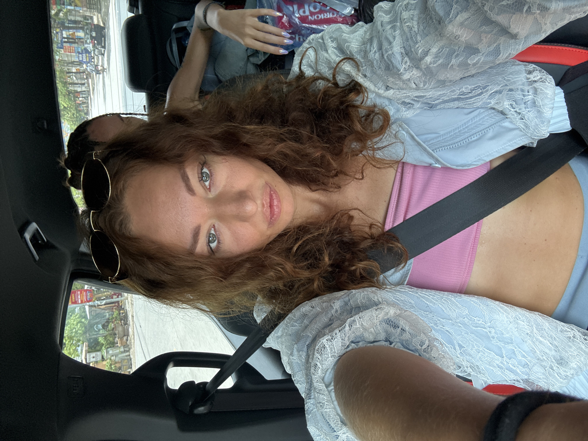 The image presents a selfie taken from inside a vehicle, depicting a scene in Trung Tam Ward, Vietnam. In the foreground, a female subject occupies the front passenger seat, oriented towards the camera. She has long, curly, reddish-brown hair with dark sunglasses positioned on top of her head. Her light-colored eyes directly engage the viewer, and her lips are slightly pursed. She is dressed in a pink ribbed bandeau top, overlaid with a light blue long-sleeved garment featuring intricate white lace patterns. A black seatbelt diagonally crosses her torso. A dark wristband is visible on her left wrist, which extends to hold the camera.
In the background, behind the primary subject, a second individual is seated in the rear of the vehicle. This person's dark, braided hair is partially visible, along with their right arm and hand. The hand, adorned with iridescent purple nail polish, rests on either the back of the front seat or on items in the rear. The vehicle's interior features a dark headliner and black seat components. A red and white plastic bag with the partial text "PRION CR" is discernible among other items in the back seat.
Through the vehicle's windows, an urban street environment is visible. The front passenger window reveals a paved road lined with trees, multiple roadside signs on poles, and several potted plants or decorative elements. The rear window offers a view of a similar streetscape, including additional trees, more unreadable street signs, a dark passenger car, and a motorcycle. The scene is illuminated by daylight.