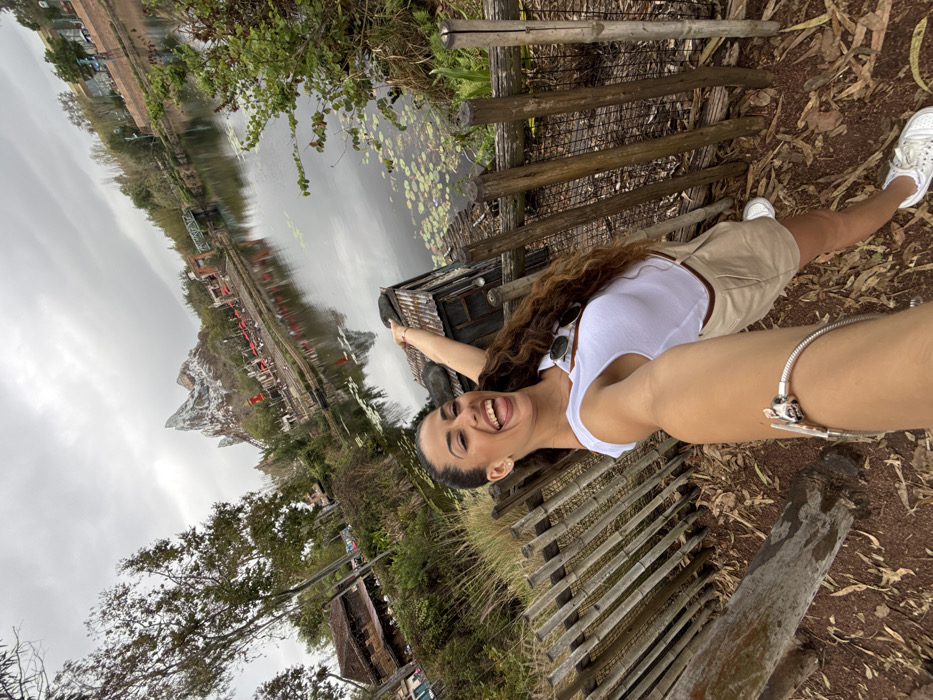 A female subject, appearing to be in her 20s or 30s with long, dark, curly hair, is captured in a selfie. She wears a white sleeveless top, light tan shorts, and white sneakers. Her left arm is extended towards the camera, revealing a silver charm bracelet on her wrist. She is smiling broadly with her mouth open and tongue slightly extended. Her right arm is raised and pointed backward towards the background.
Immediately behind her is a rustic fence constructed from vertical wooden logs and horizontal poles. The ground beneath her is reddish-brown earth with scattered leaves. To her right, behind the fence, is a small, weathered wooden structure with a rusty corrugated metal roof.
Beyond the fence and structure, a body of still water with lily pads covers the midground, reflecting the overcast sky. On the far side of the water, a prominent, artificial mountain peak, identifiable as Expedition Everest, dominates the background. Associated structures, including buildings with red roofs, lush trees, and a green metal truss bridge spanning the water, are also visible. The sky is uniformly overcast.
The scene is located at Disney's Animal Kingdom theme park in Bay Lake, United States.
