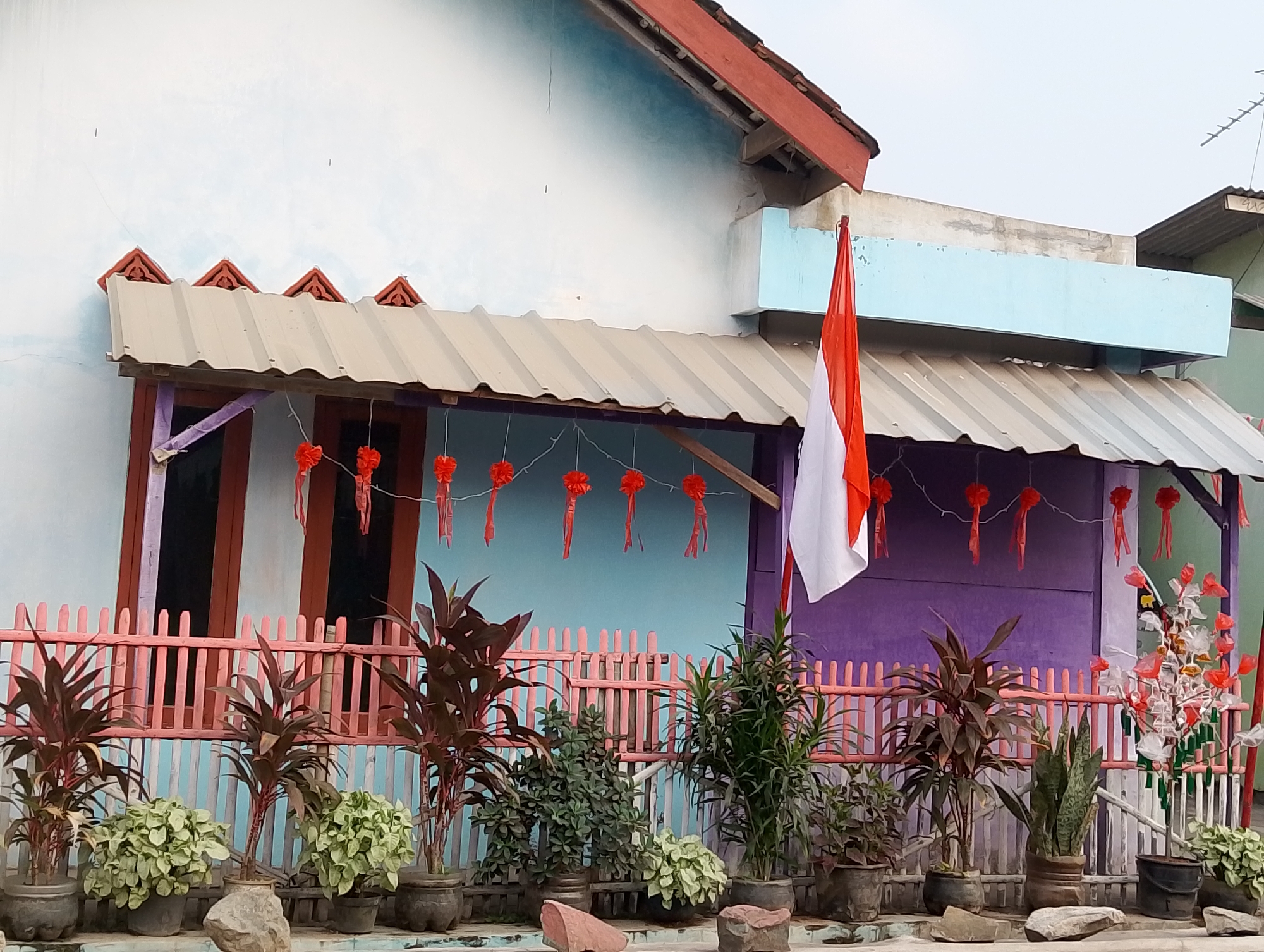 The image shows the facade of a house decorated for a national celebration, likely in Karawang, Indonesia, given the Indonesian flag displayed. The house has a light blue wall, a corrugated metal roof, and a red-tiled overhang. A pink wooden fence runs horizontally in front of the house, behind which several potted plants are arranged. A prominent Indonesian flag, red and white, is affixed to a pole to the right of the entrance. Red decorative streamers are strung across the overhang. The scene is outdoors, with the sky visible in the background. There are no people visible in the image.