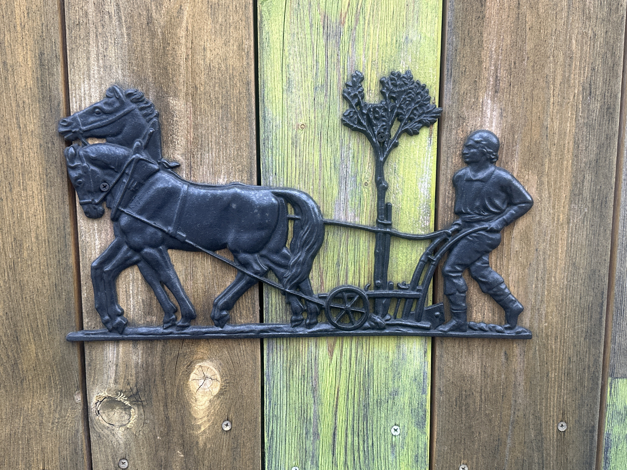 A dark, cast iron decorative plaque depicts a scene of a man with a horse-drawn cart. The plaque is mounted on a wooden fence. The fence is composed of vertical planks, with some planks painted a bright, weathered green and others a natural wood color. The plaque features two horses in the foreground, harnessed together and appearing to pull a cart. The cart is a simple, two-wheeled design, with a small tree attached to its side. Behind the cart, a man is shown in profile, facing left, holding the cart's handles as if he is guiding it. The man is wearing simple clothing, including trousers and a shirt, and appears to be walking. The plaque is secured to the fence with visible screws. The overall scene suggests a rustic or historical depiction of farm work or transportation.