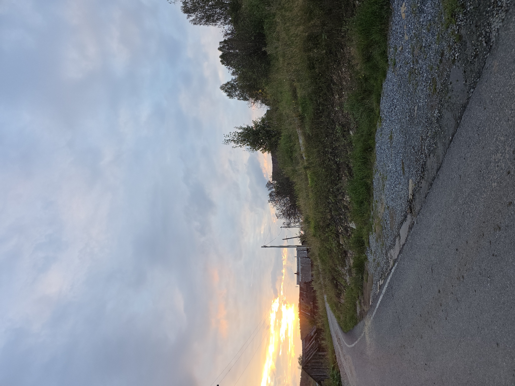 The image shows a rural road winding through a landscape with a dramatic sunset sky. The road is paved and appears to be a two-lane rural road with a gravel shoulder. On the left side of the road, there are several dilapidated wooden structures that appear to be old farm buildings or abandoned houses, some with rusted corrugated metal roofs. The buildings are set against a backdrop of trees and the glowing sunset. The right side of the road slopes upwards into a grassy hillside dotted with trees. The sky is filled with a mix of clouds, with streaks of orange and pink from the setting sun visible on the left horizon. Power lines and poles are also visible, stretching across the scene. The overall impression is one of a remote, rural setting at dusk. No people are visible in the image.