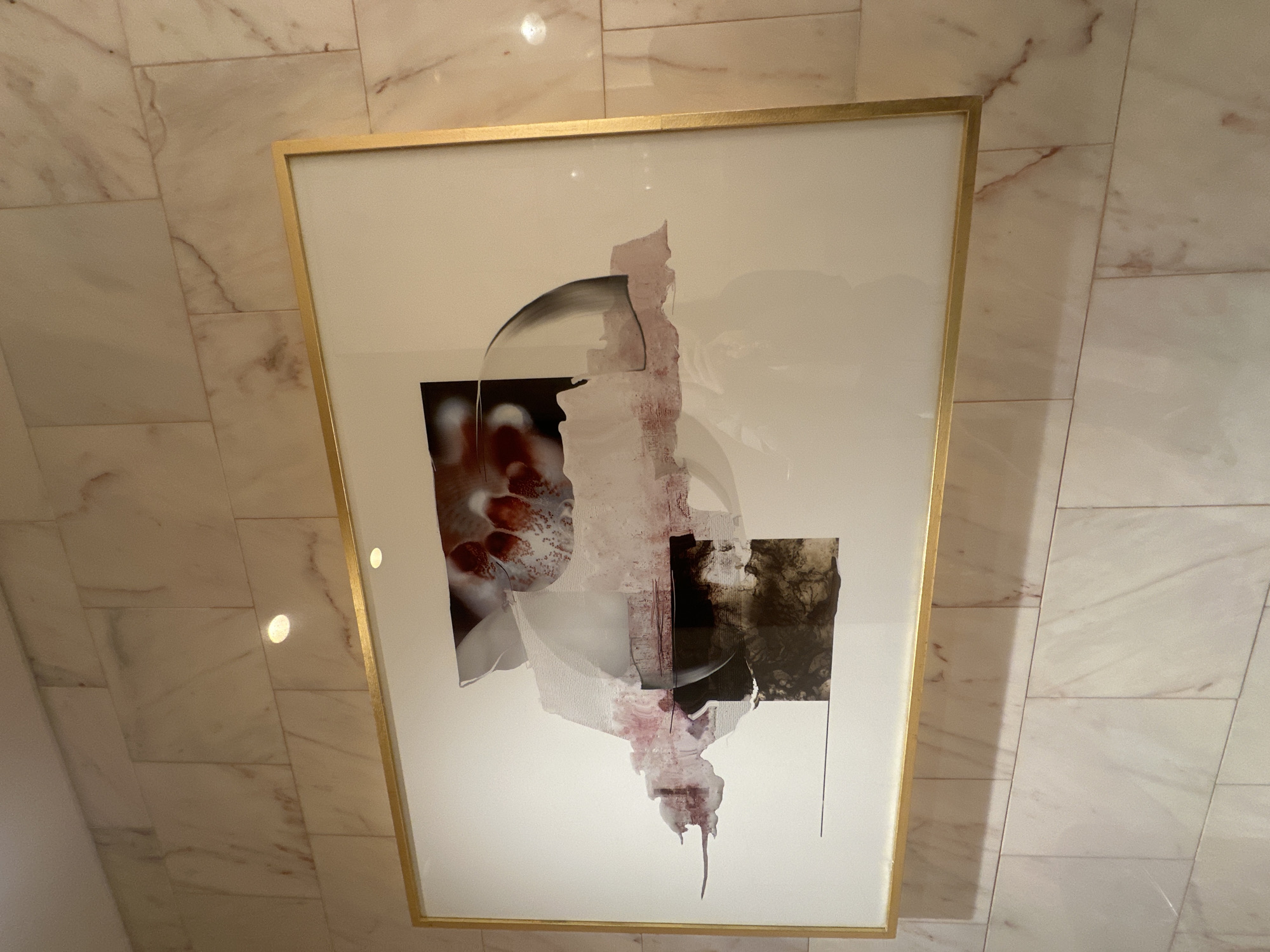 The image shows a framed abstract artwork hanging on a wall covered in marble tiles. The artwork features a white background with overlapping abstract shapes in various colors, including red, gray, and brown. The frame is gold. The location is not discernible from the image.