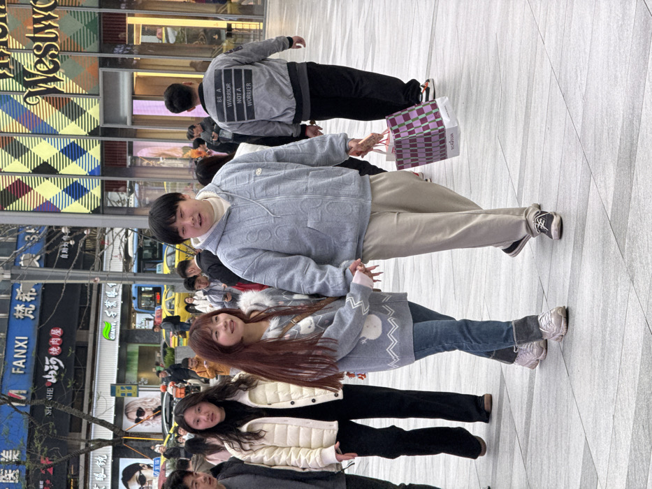 The image depicts a bustling urban pedestrian area in Liangjiang, China. The ground is paved with light gray rectangular and square tiles.
In the foreground, an adult East Asian couple walks forward, holding hands. The female, on the left, has long reddish-brown hair and wears a light gray sweater with white patterns, blue jeans with rolled cuffs, light-colored athletic shoes, and a light-colored crossbody bag. Her fingernails are long and pointed. The male, on the right, has short dark hair and wears a light gray hooded zipper jacket with embossed patterns over a white high-neck shirt, light beige sweatpants, and dark sneakers. He carries a purple and pink checkered "YOOGRAN" shopping bag in his right hand.
To the immediate left of the couple, an adult female with long dark hair walks forward, wearing an off-white padded jacket, a black turtleneck, and black wide-leg trousers. Behind the couple, on the right, an adult male walks away, wearing a gray bomber jacket with the white text "BE A WARRIOR NOT A WORRIER" printed across the back in four lines, dark pants, and white sneakers with orange accents.
Numerous other pedestrians are visible in the background, walking in various directions. Commercial buildings line the street. To the left, a building features blue signage with "FANXI" and Chinese characters, and "SHEN HE 小山河烤肉屋" (Xiaoshanhe Roasted Meat House) below, alongside large display advertisements for sunglasses. To the right, a modern building exhibits a multi-colored checkered facade in yellow, blue, and purple, with "Westwood" partially visible. In the distant background, yellow taxis and a blue public bus are present on an adjacent street.