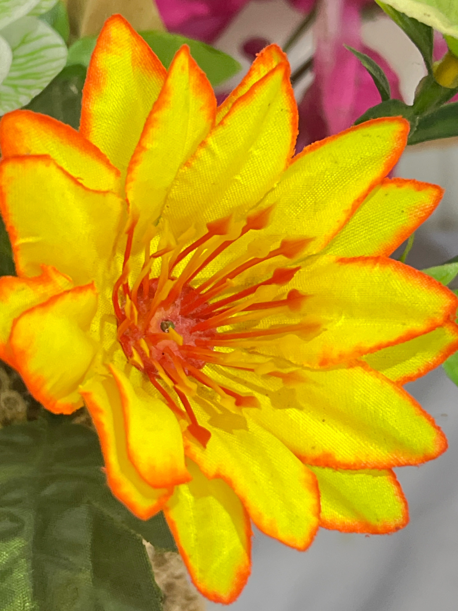 This is a close-up, eye-level, medium shot of a bright yellow and orange artificial flower. The flower has multiple layered petals that are edged in a vibrant orange. In the center of the flower, there are numerous thin, red stamens tipped with orange. The background is softly blurred, showing hints of green foliage and some pink and white shapes, suggesting other artificial flowers or decorative elements. The lighting is bright, illuminating the textures and colors of the artificial flower. There are no people or animals visible in the image. The scene does not provide any information to link it to Denpasar, Indonesia.