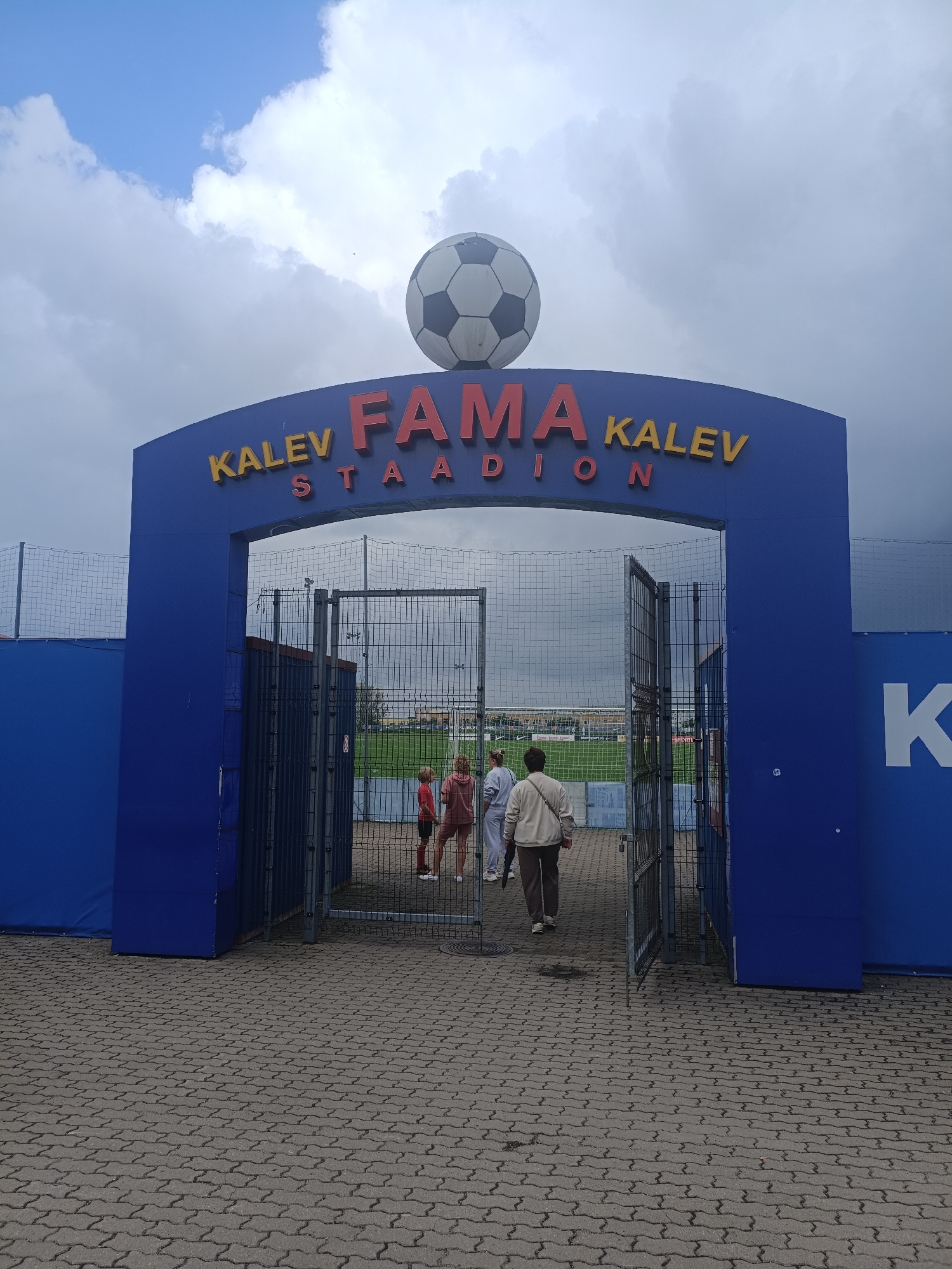 The image shows the entrance to the Kalev Fama Stadium in Narva, Estonia. The entrance is a large blue archway with the words "KALV FAMA STADION" in red and yellow letters. A large soccer ball is mounted on top of the arch. Through the open gates, a soccer field is visible, along with a few people standing near the entrance. The sky is cloudy.