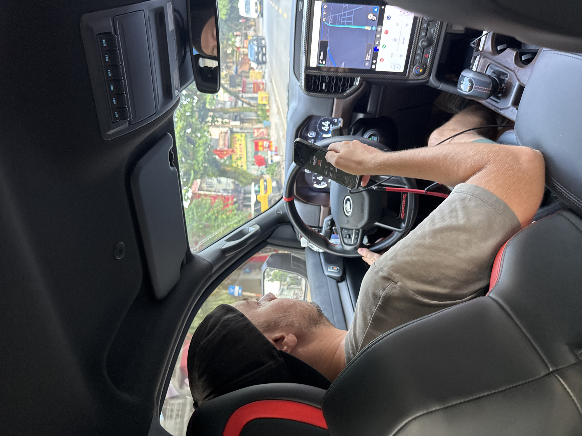 A view from inside a vehicle shows a man in the driver's seat, seemingly operating or engaging with a smartphone while stationary or moving slowly in Vị Xuyên Commune, Vietnam. He is seen from the side and slightly from behind, wearing a gray t-shirt and khaki shorts, with a black cap covering his head, leaning back against the seat's headrest. His left hand holds a smartphone displaying an application with visible text "ALERTS" and "REAR TV", while his right hand rests on the steering wheel, which features a "FORD" logo.
The car's interior is modern, with a large, vertically oriented central infotainment screen displaying a navigation map that shows a route, estimated time (e.g., "25 min"), and various app icons, including "Q.Y.O." in the top right. A digital instrument cluster behind the steering wheel shows the number "44". An overhead console has a row of six illuminated buttons labeled "AUX 1" through "AUX 6". The seats are black leather with red accents, and the gear shifter is visible on the center console.
Through the windshield, a busy street scene unfolds under bright daylight. Buildings, storefronts with indistinct signs, lush trees, and other vehicles, including a white van and a dark car, are visible, suggesting an urban or semi-urban environment. The overall impression is of a driver taking a break or navigating in traffic, using his phone.