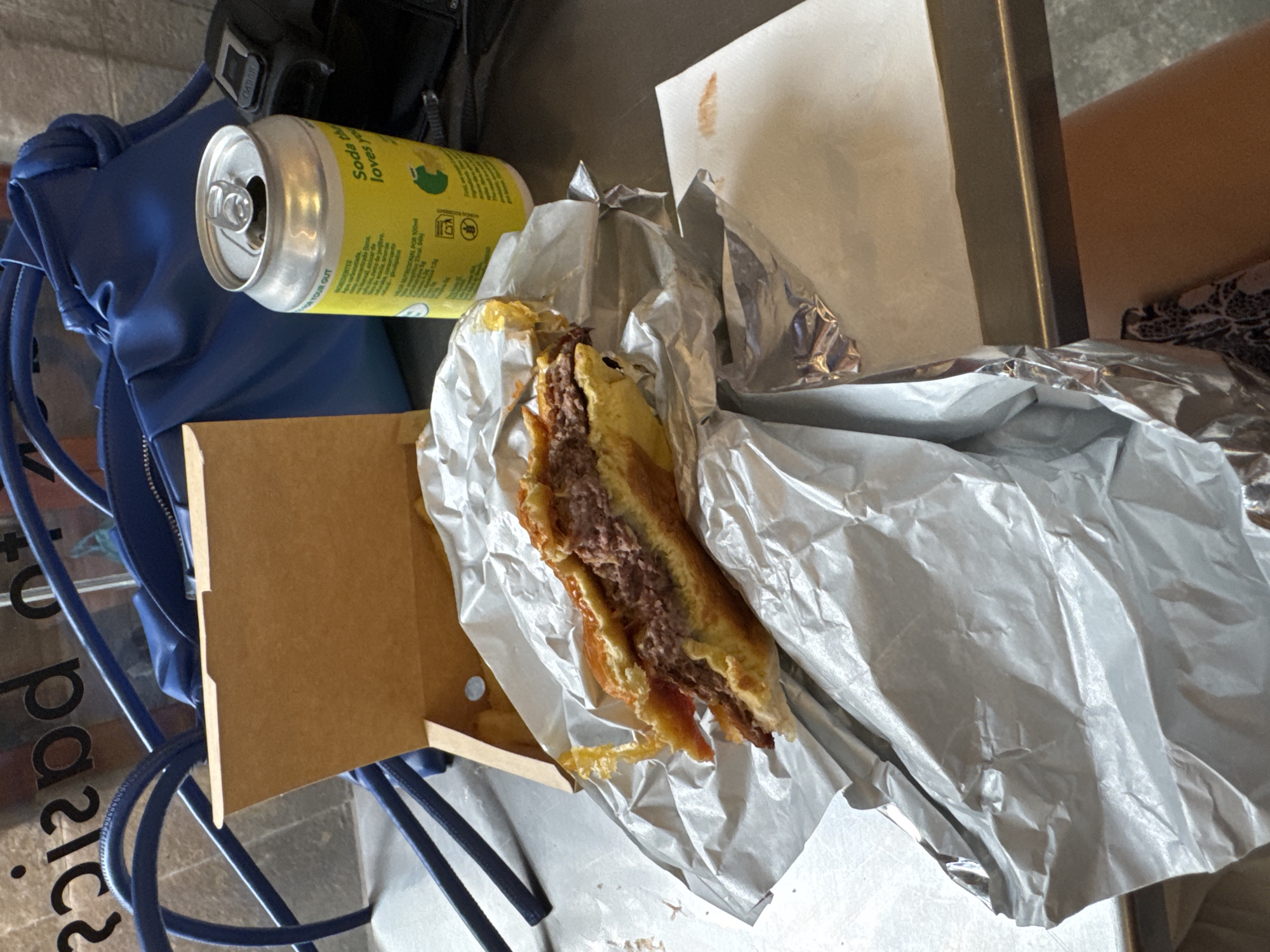 This photo captures a casual scene with an assortment of items. There is a partially eaten burger nestled in silver foil inside a cardboard container, accompanied by a can of "Soda" beverage. A blue backpack is visible, and the scene appears to be set on or near a metal chair. There is also a book or paper product with a stained mark on it. The location is Barcelona, Spain as indicated by the letters of the city in the background.