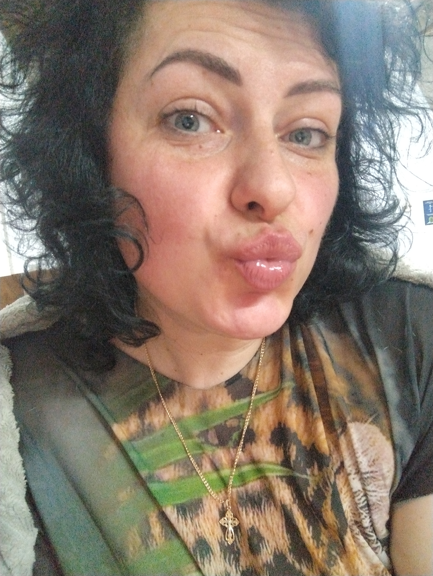 A woman with fair skin, dark curly hair, and light-colored eyes is seen in a close-up shot. She is wearing a patterned shirt with a print resembling tiger stripes and a gold necklace with a cross pendant. Her lips are pursed in a kissing expression. The location is unspecified and no information indicates it is Grishanki, Russia.