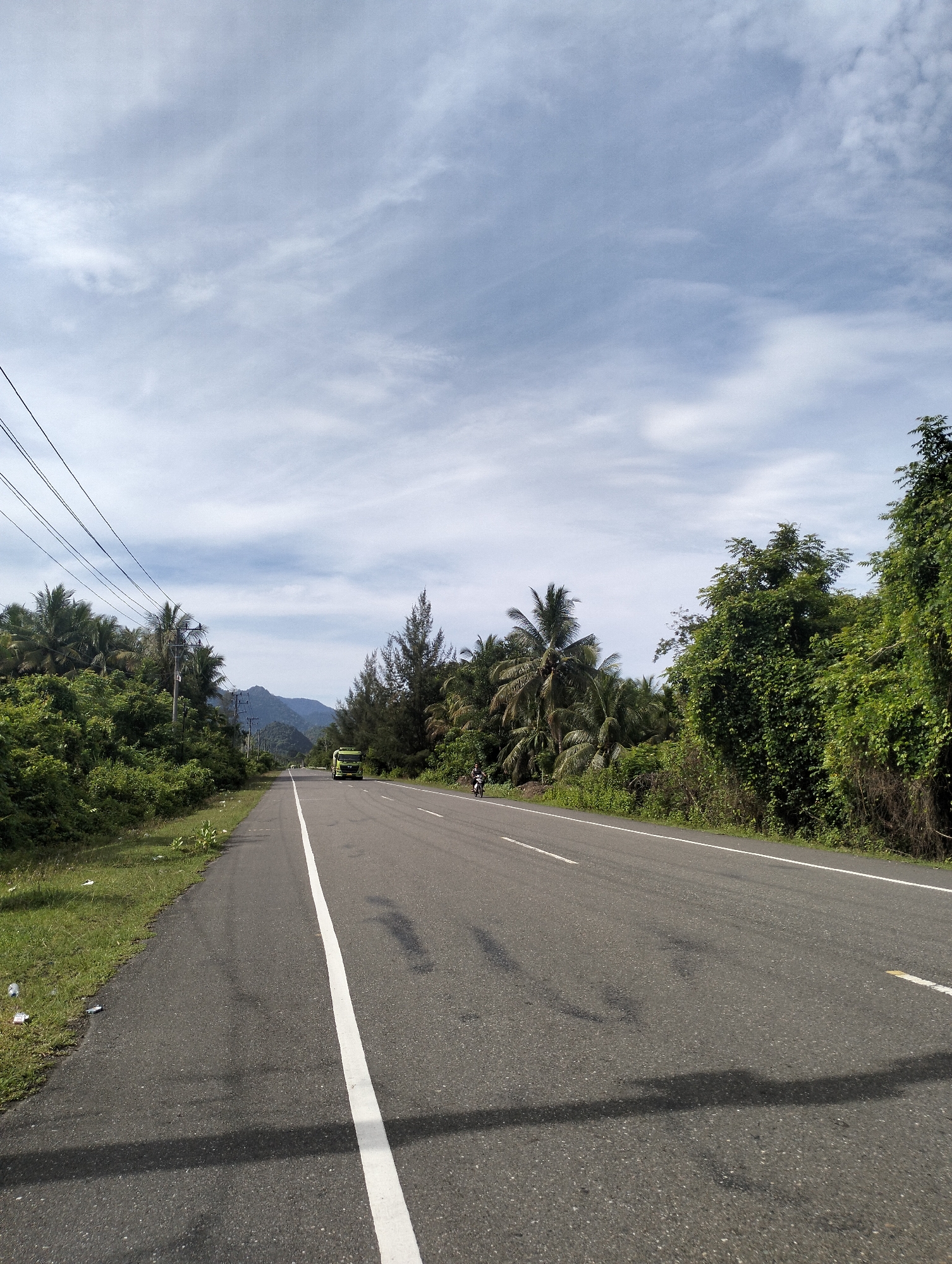 The image depicts a straight stretch of asphalt road in Meunasahmesjid, Indonesia. A solid white line divides the road. Vegetation flanks both sides of the road, transitioning into dense greenery. In the distance, a green truck and a motorbike are visible on the road. Overhanging power lines are present on the left side. The sky is overcast with patterned clouds. Mountains are subtly visible in the distance, beyond the trees. Debris is visible on the left side of the road.