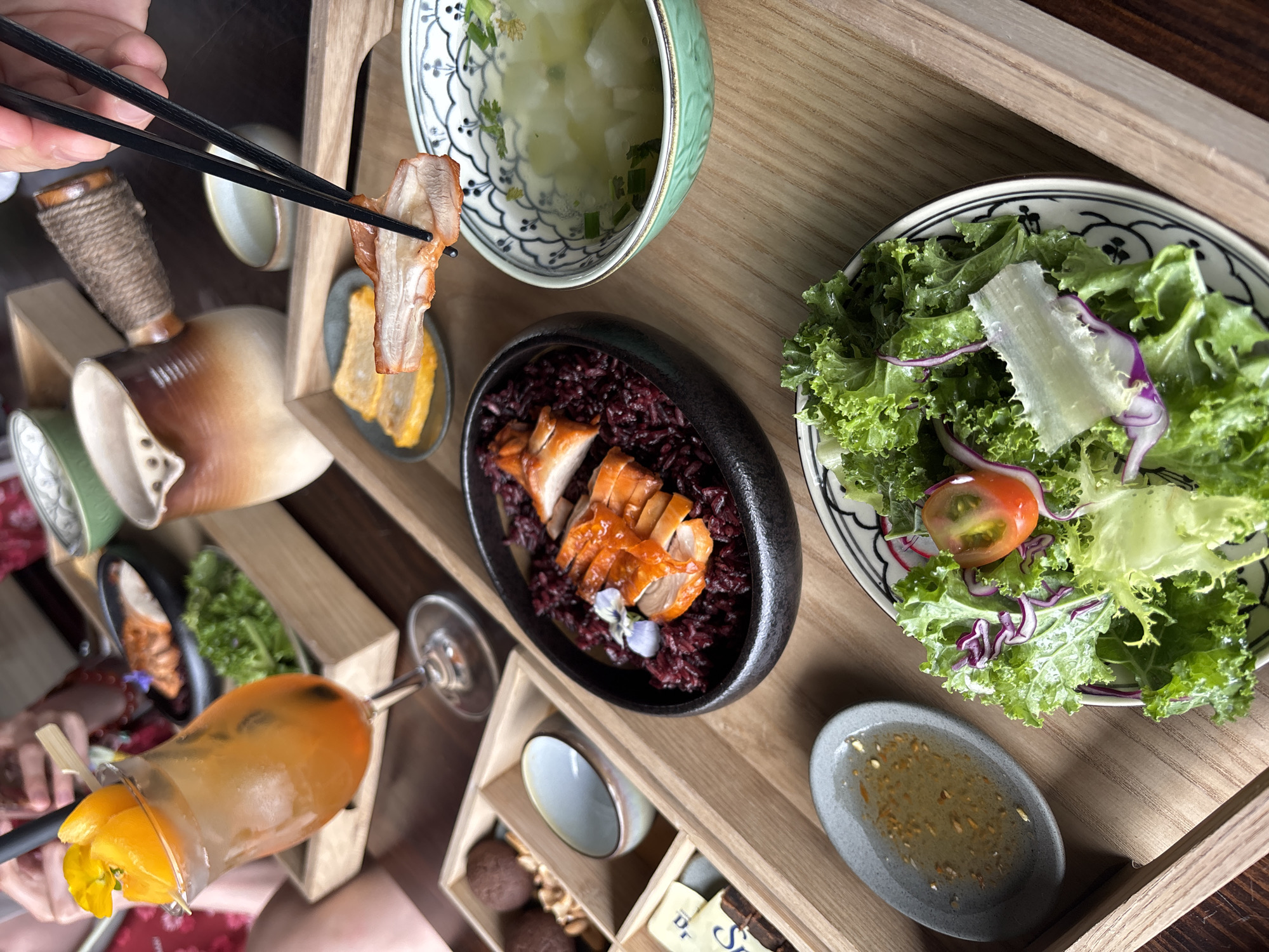 A close-up, overhead shot captures a wooden tray filled with a meal, likely in Da Lat, Vietnam, given the presence of traditional Vietnamese cuisine elements. In the foreground, a salad with vibrant green lettuce, thinly sliced red cabbage, and a halved cherry tomato is presented on a patterned plate. To its left, a small gray dish holds a light-colored dressing with flecks of herbs and spices.
Dominating the center of the tray is a black bowl filled with dark purple rice, topped with slices of roasted duck or pork, garnished with a delicate purple flower. To the right of this dish, a green and white patterned bowl contains a clear soup with pieces of white vegetables and green herbs.
Further back, a person's hand is using black chopsticks to pick up a piece of roasted meat. Behind this, a beverage in a tall glass with an orange hue and garnished with orange slices and a yellow flower sits on the tray. Another small dish with what appears to be two pieces of fried food is visible to the left of the meat being picked up.
The wooden tray itself is partially seen, suggesting it rests on a darker surface, likely a table. In the background, a rustic ceramic pot with a rope-wrapped handle is visible, alongside other small dishes, and hints of other diners and a wine glass. The overall scene suggests a well-presented, individual meal with several components.