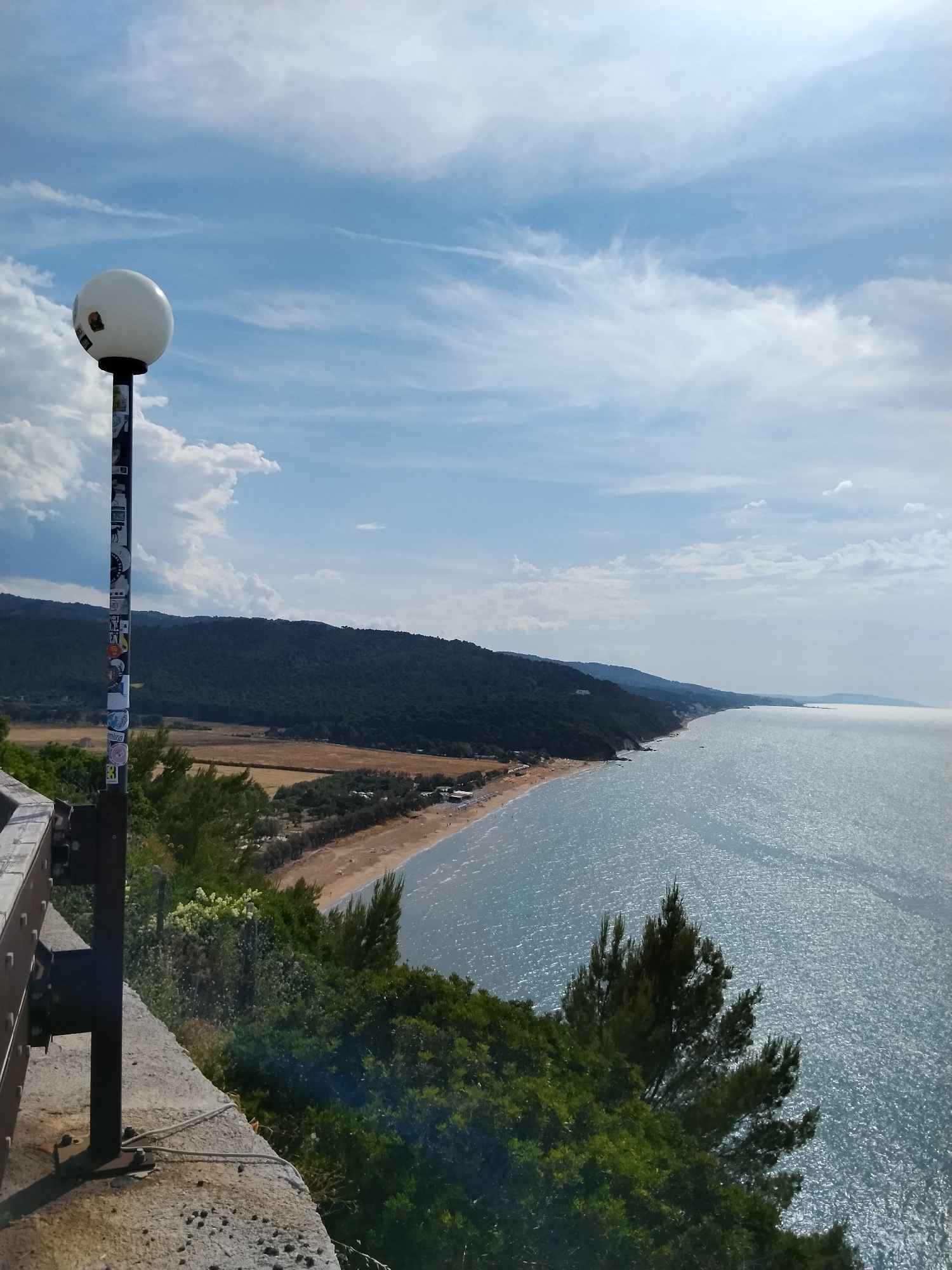 This detailed photograph captures a panoramic coastal landscape from a high vantage point in Vico del Gargano, Italy. In the immediate foreground on the left, a dark metal lamppost, adorned with numerous multi-colored stickers, rises from a concrete ledge. The lamppost is topped by a large white spherical light fixture. A metal guardrail is partially visible on the far left of the concrete structure. Lush green vegetation, including evergreen trees, lines the steep slope below the vantage point and extends along the coastline.
The midground reveals a long, sandy beach populated by small, indistinct figures and dotted with beach umbrellas. Inland from the beach, a strip of lighter-colored terrain, possibly fields, separates it from a series of densely forested hills and mountains that run parallel to the coast.
To the right, the glistening blue-green water of the Adriatic Sea stretches to the horizon, reflecting bright sunlight across its rippled surface. The coastline extends into the distance, where the forested hills meet the sea. The sky above is a bright blue with scattered white clouds, ranging from wispy cirrus to more defined cumulus formations, indicating a sunny day.