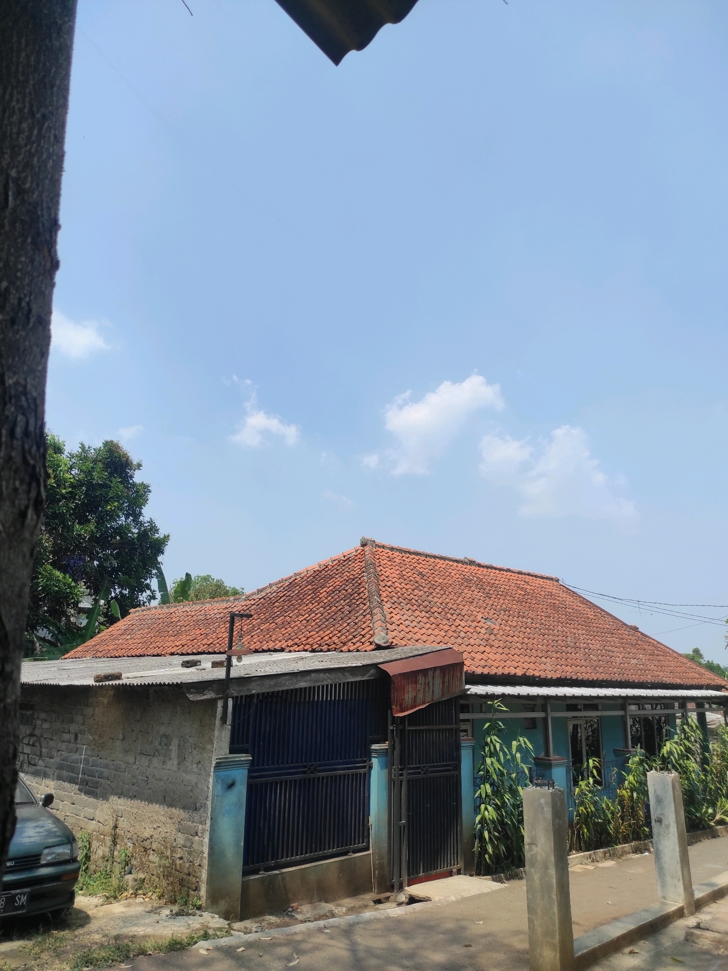 A residential house in Padalarang, Indonesia, is shown under a clear blue sky with sparse white clouds. The house features a prominent red tile roof with a slight slope. The walls of the house are painted a light blue. A dark metal fence runs along the front of the property, and a section of this fence is open. To the left of the house, there is a gray brick wall and a portion of a dark-colored car is visible parked on the street. The street appears to be paved. Green trees and vegetation are present in the background and to the side of the house. Two unfinished concrete posts stand in the foreground, on the right side of the image. The overall scene suggests a typical residential area during daylight hours.