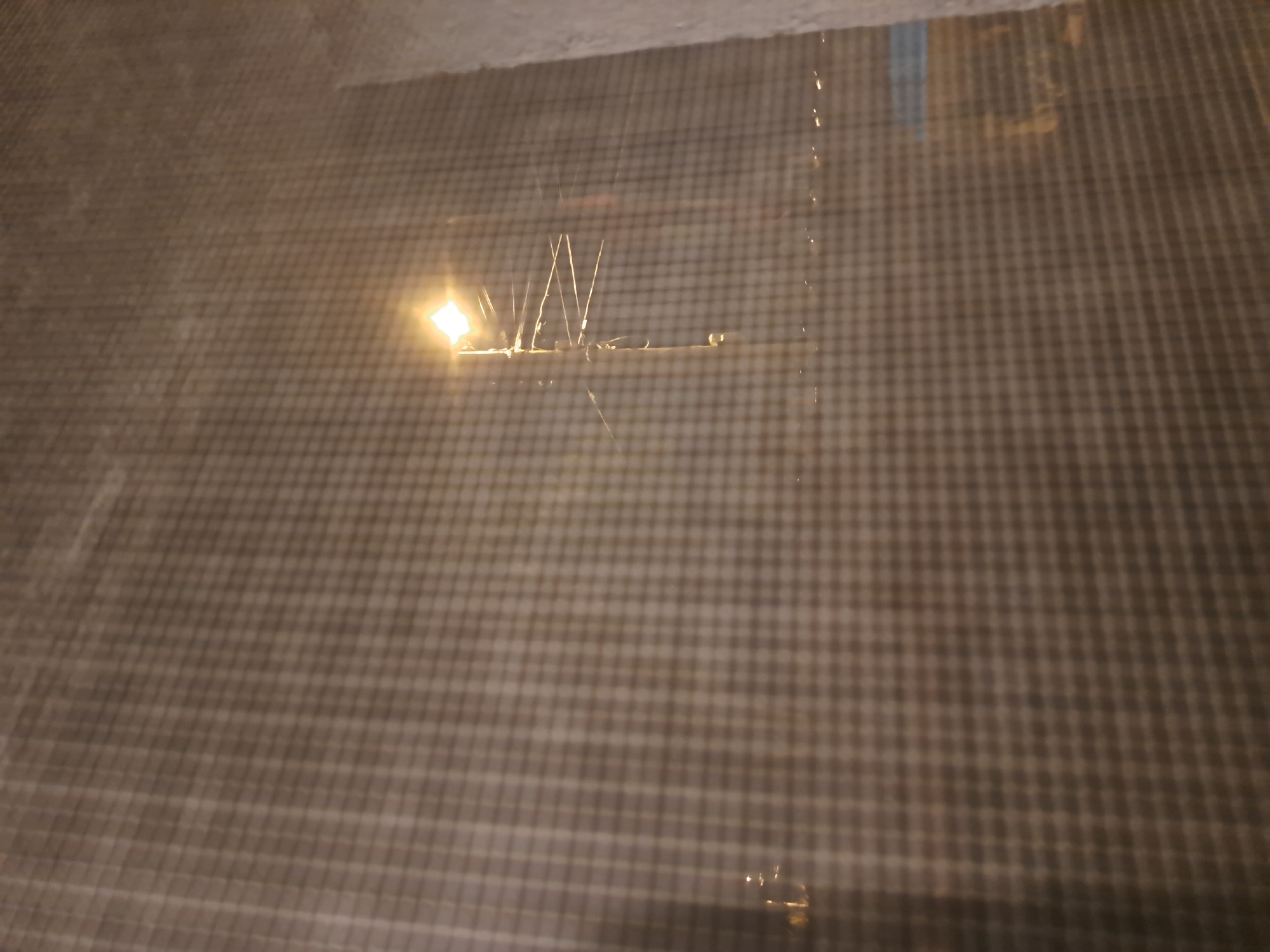 The image shows a window with a grid pattern. Outside the window, there's a light source, possibly a streetlamp or building light. Above the light source, there are bird spikes installed on a ledge. The scene appears to be at night or in a low-light environment. I do not have enough information to pinpoint the location in Negovan, Bulgaria.