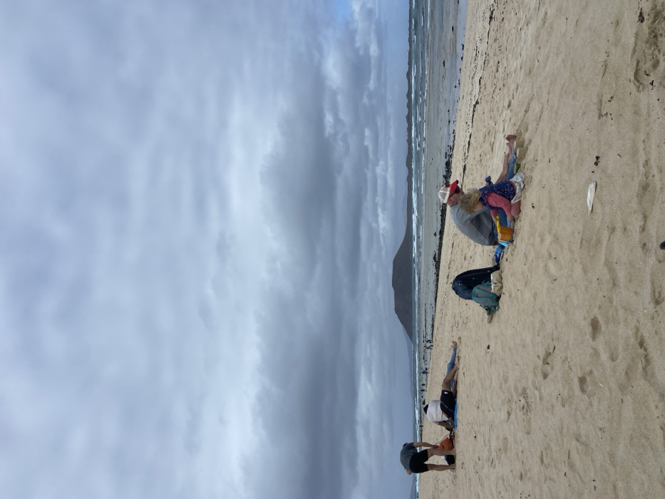 The image shows a beach scene in Corralejo, Fuerteventura, Spain (latitude: 28.724, longitude: -13.842). The sky is overcast with white clouds. The beach is a wide expanse of light-colored sand. Three people are sitting on the sand, while several more are in the distance. The person closest to the viewer is wearing a blue shirt, blue shorts, and is bending over. Next to them is another person, wearing a red shirt, black shorts, and a white hat. They are sitting on a blue and black backpack. The person furthest from the viewer is wearing a red shirt and black shorts. The scene is peaceful and tranquil.