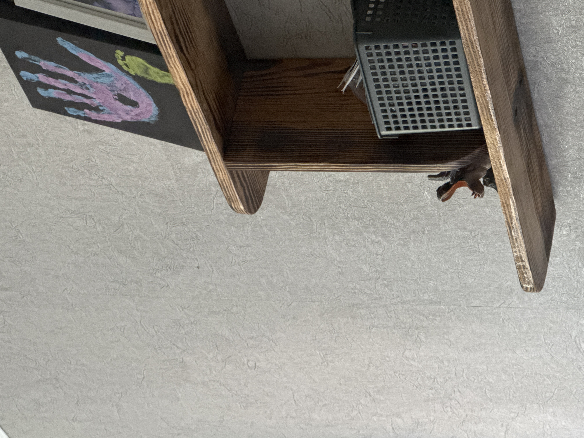 The image shows a wooden shelf with a black framed artwork of a handprint and footprint on the left. On the right, a gray plastic container with a grid pattern sits on the shelf. A toy dinosaur peeks out from under the shelf on the right side. The background is a textured light gray wall. The lighting suggests it is daytime.