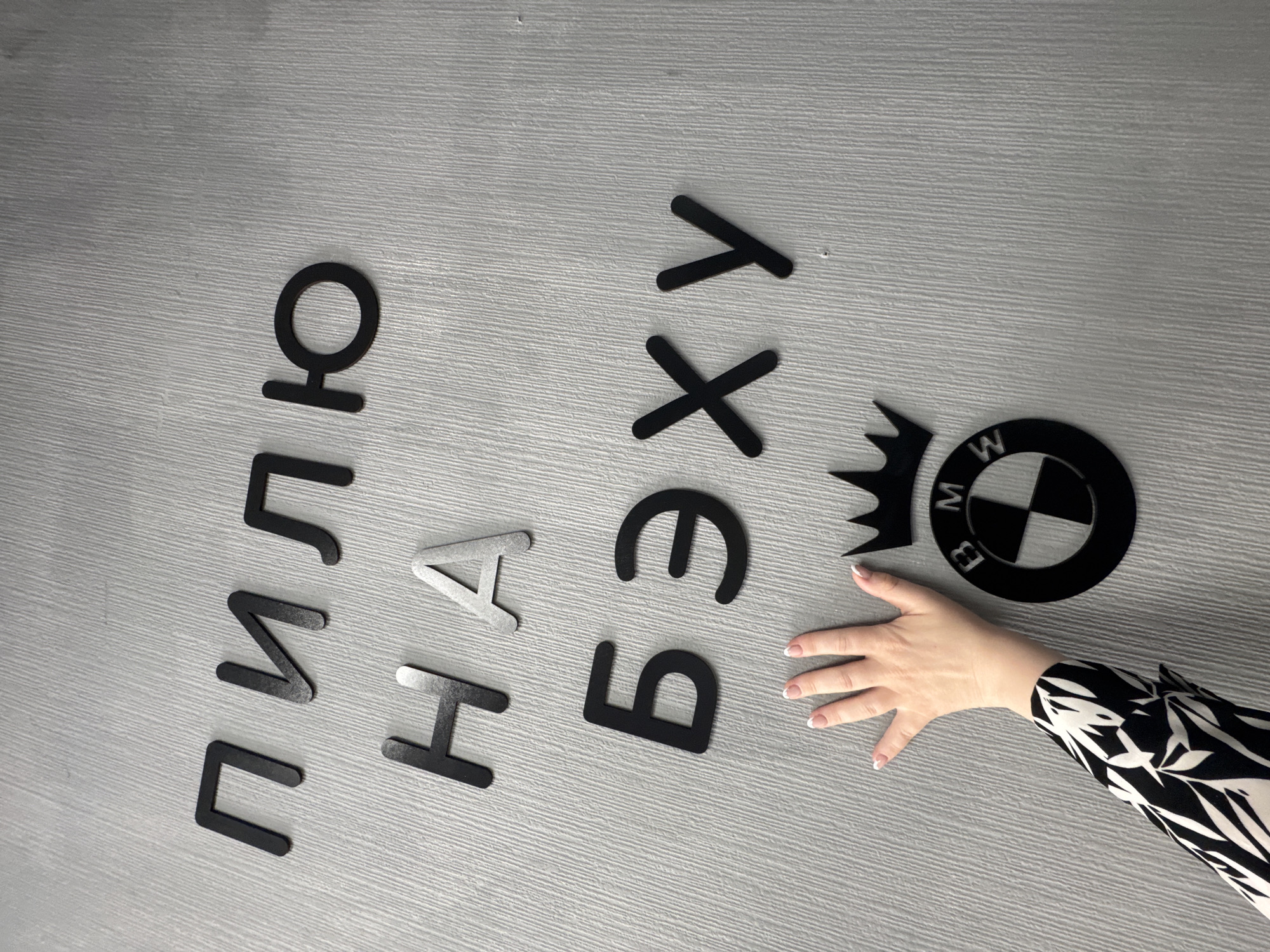 A high-angle medium shot shows the black cutout Cyrillic letters and a BMW logo arranged on a grey surface. From left to right, the letters spell "Пильно На Взгляд". A woman's hand with a black and white patterned sleeve reaches towards the BMW logo. The shot may be taken in Chaikovsky, Russia, but there is no evidence to confirm it in the picture.