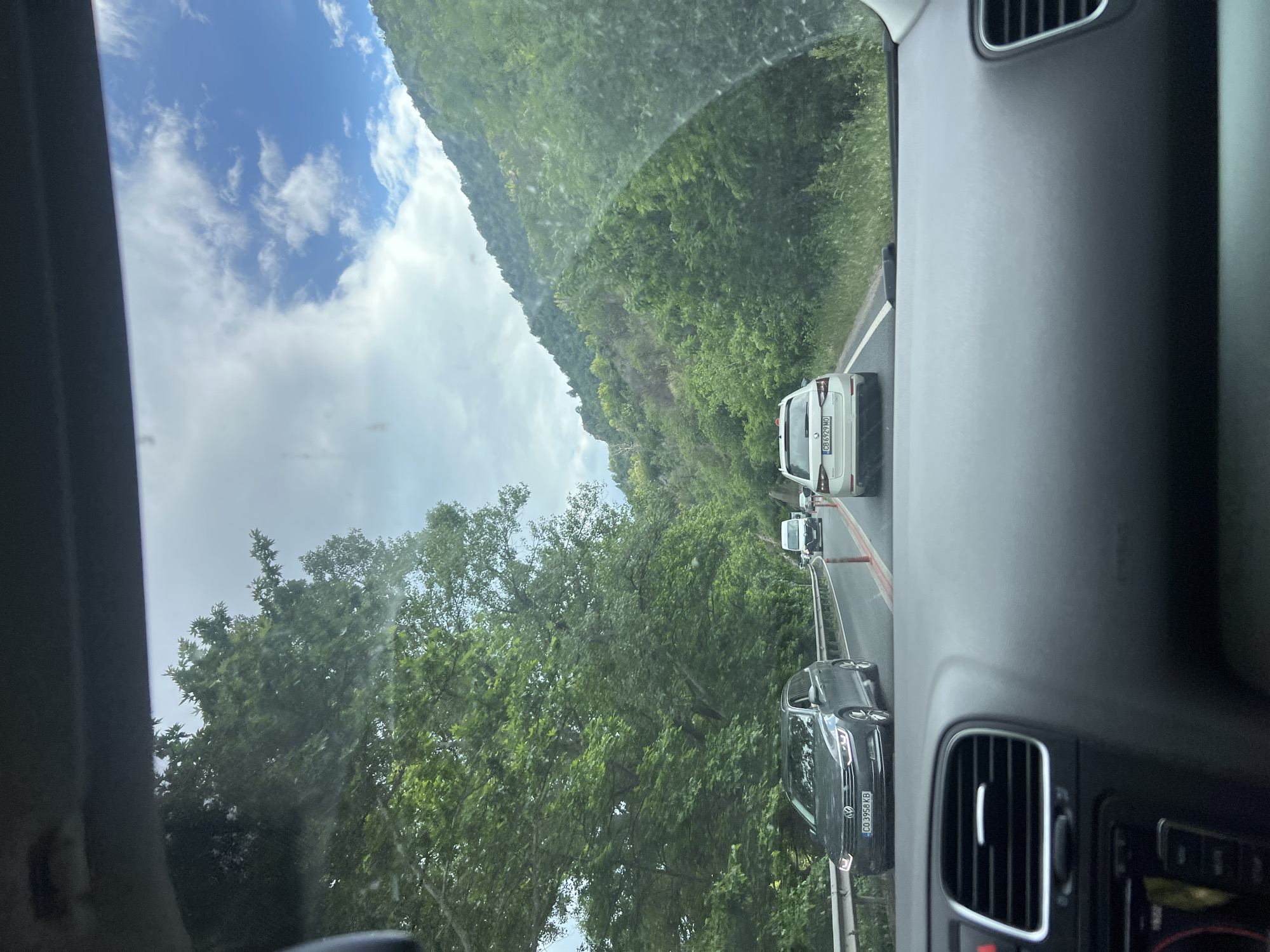 The image, taken from inside a vehicle, shows a roadway in Stara Kresna, Bulgaria. Three cars are visible in front of the camera car, with the last car closest being dark gray, the second being a white van with a visible "CB 629 MO" license plate, and the furthest a white hatchback. A green mountainside rises on the right side of the road, while trees line the opposite side, framing the partly cloudy sky. The camera is pointed slightly upwards and slightly towards the left. Parts of the dashboard and air vents of the vehicle are visible.