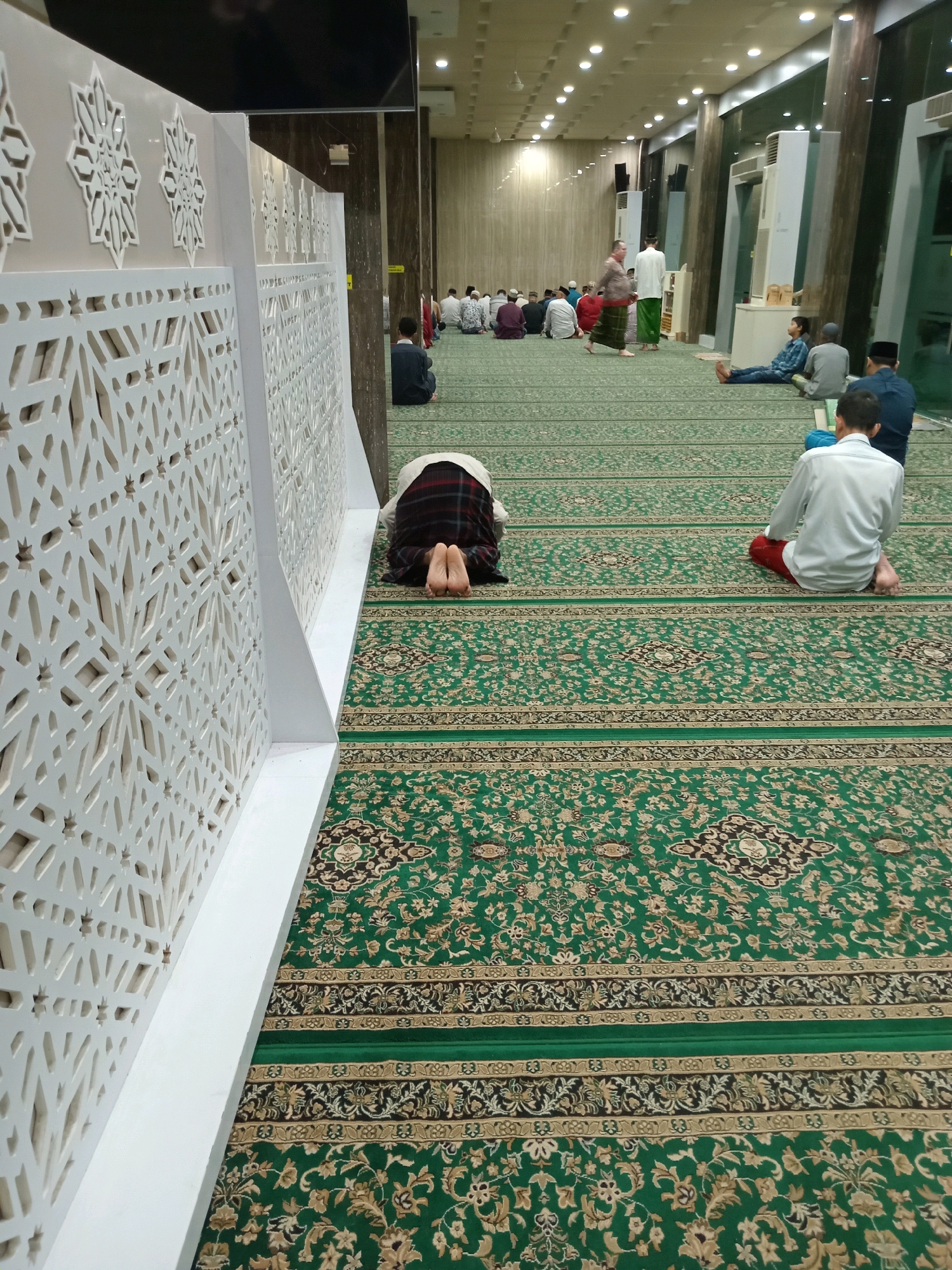 A long shot shows the interior of a mosque in Randuagung, Indonesia, with a green carpet covering the floor. The carpet is decorated with intricate patterns in brown and gold. In the foreground, a person is seen bowing in prayer, with their head and arms resting on the carpet. Behind this person, several other individuals are also seated on the carpet, facing the same direction, indicating they are also engaged in prayer. The mosque's interior features white decorative panels along the left wall, adorned with geometric patterns. The ceiling is illuminated by numerous lights. In the background, more people are seated on the carpet, and some are walking. The scene suggests a communal prayer gathering within the mosque.