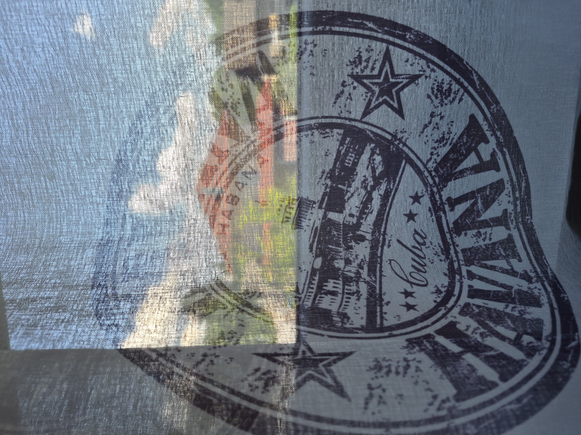 This is a close-up shot of a sheer, light blue fabric with a printed design. The fabric is draped, creating folds and shadows. A distinct graphic is printed on the right side of the fabric, appearing as a dark, distressed stamp or seal. It features a star at the top, a circular border, and stylized lettering. Partially visible within the circle are shapes that resemble buildings or a cityscape, rendered in a dark, textured print. The word "Cuba" is clearly legible in cursive script, accompanied by two small stars. The fabric's transparency allows the background to be visible, which appears to be an outdoor scene. The background shows a bright sky with wispy clouds and what looks like the reflection or impression of foliage and possibly some structures with red and orange elements, suggesting a sunny day. The light filtering through the sheer fabric casts a diffuse glow on both the fabric and the background scene. There is no visible text in the background scene. The overall impression is of a graphic design viewed through a translucent veil, with the natural light and background adding a layered depth to the image.