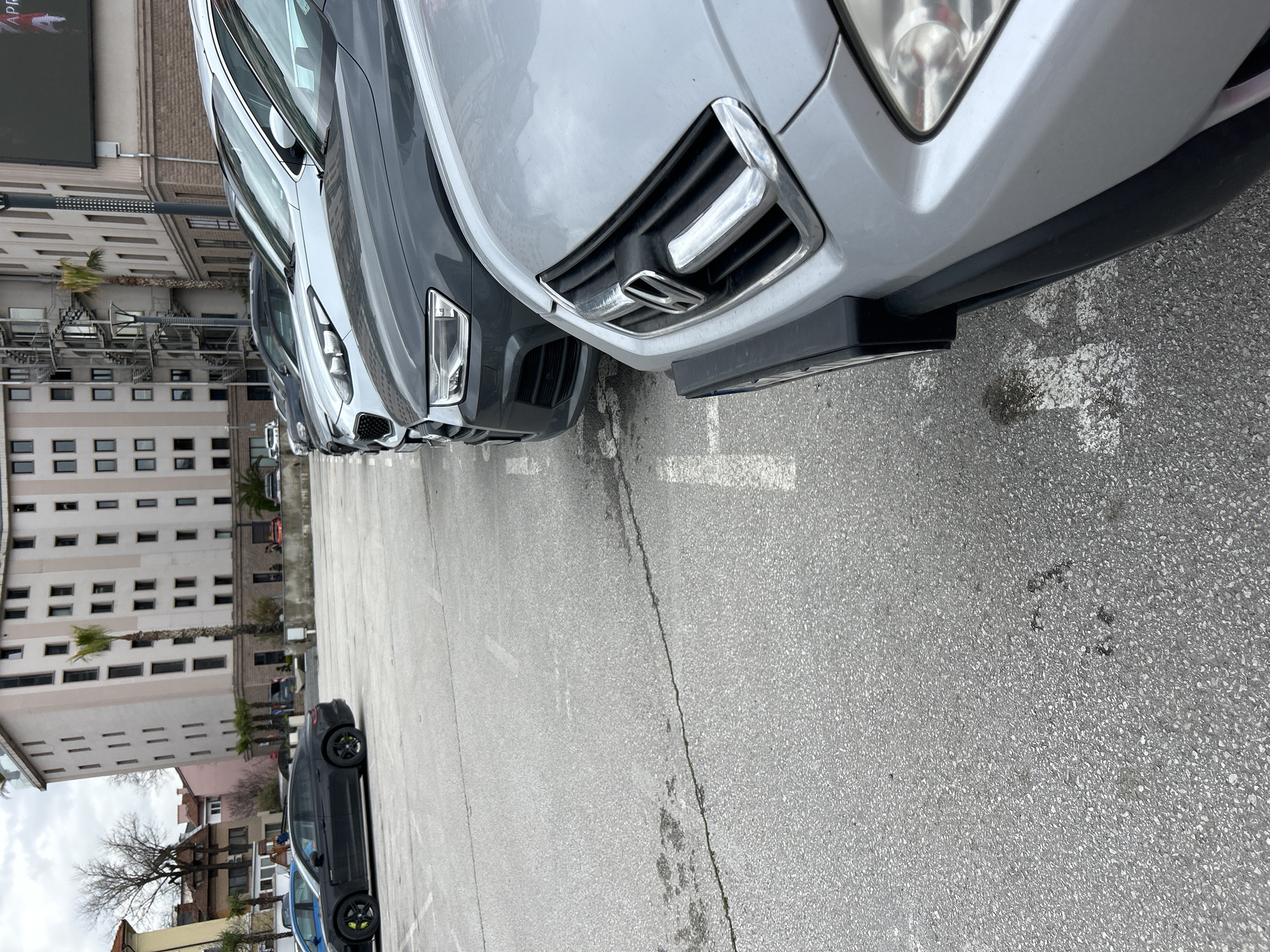 The image depicts a street scene in Plovdiv, Bulgaria. A row of parked cars, including gray and silver vehicles, dominates the foreground. The camera is positioned low, giving a close-up view of the vehicles' front ends, including details like headlights and grilles. In the background, a multi-story building with numerous windows and possibly fire escapes is visible. Also, there are some bare trees visible with no leaves on them. Further back, a few additional cars are seen along the street. The parking lot is paved and marked with white lines. The overall atmosphere is mundane and urban.