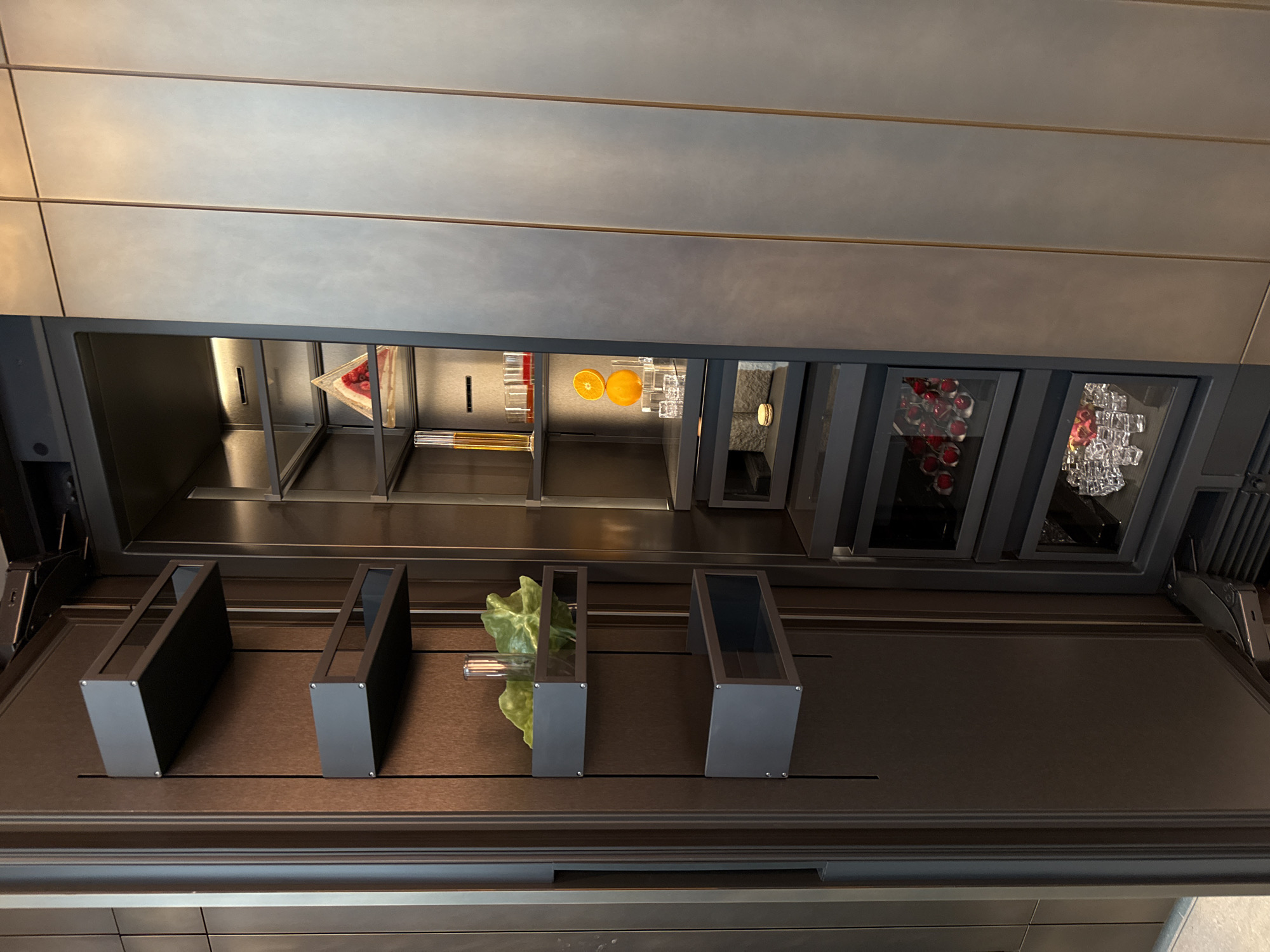 The image depicts an overhead bar setup, likely in a modern establishment, displaying various ingredients and glassware. The bar features built-in compartments holding fruit slices (oranges and berries), bottles, and an assortment of cocktail glasses. In the foreground are dark containers, one of which holds green leaves and possibly a straw. The bar appears to be well-lit and meticulously organized. The exact location in Milan, Italy is not discernible from the image.