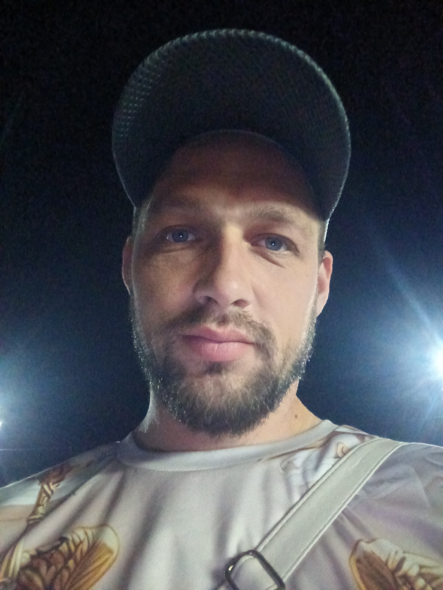 This is a selfie taken outdoors at night in the city of Balqaş, Kazakhstan. The subject is a man with blue eyes, a beard, and he is wearing a black baseball cap and a t-shirt with a graphic print. A white strap from a bag is visible across his shoulder. The background is dark, with some blurred lights, suggesting artificial lighting.