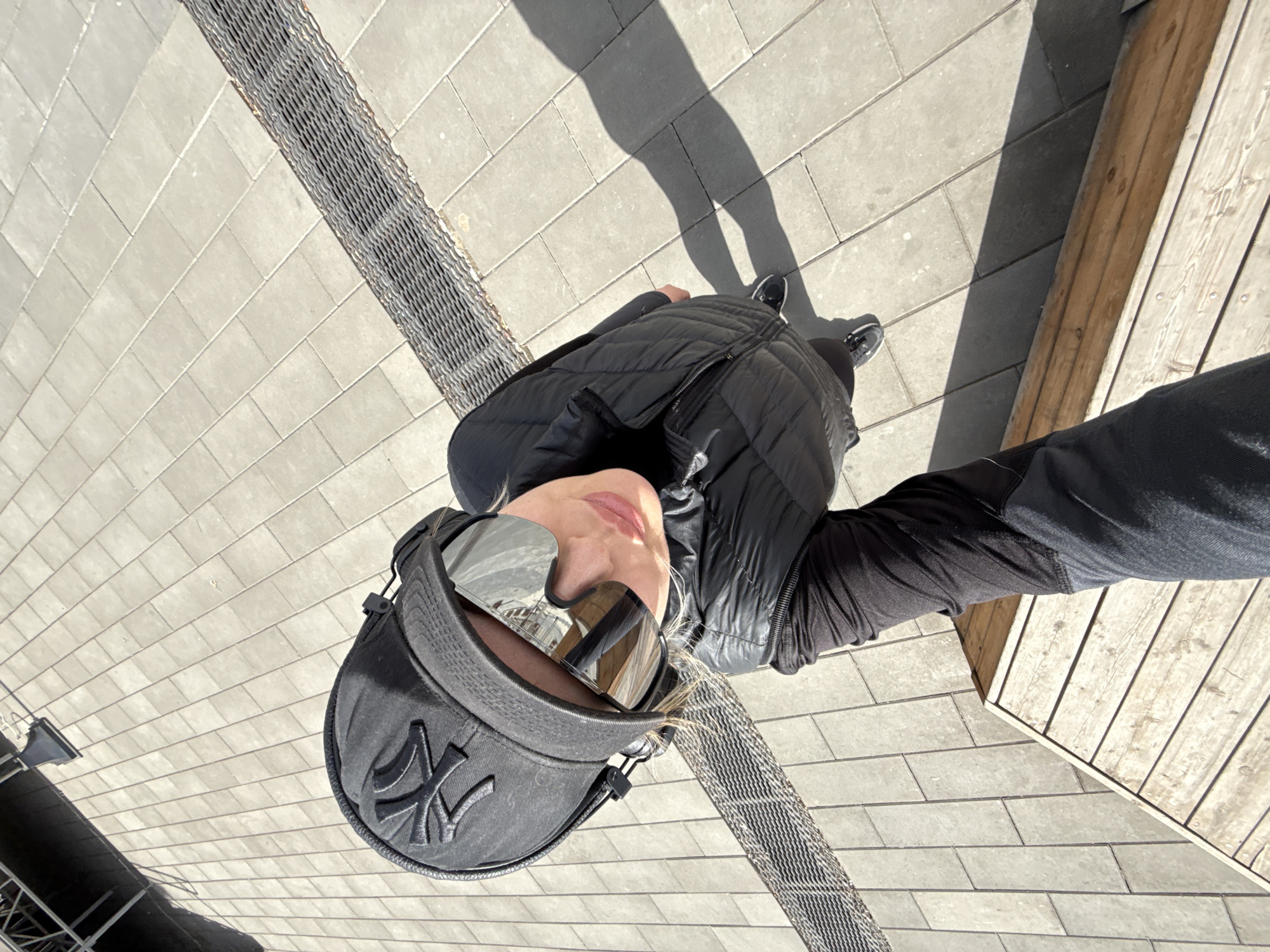 A person is seen from a high-angle perspective on a paved surface with alternating sections of brickwork and metallic grids. The individual is wearing a black baseball cap, reflective sunglasses, a black puffer vest, and dark-colored shoes. Their shadow is cast on the pavement behind them. In the right upper quadrant, a wooden structure is visible. The location cannot be determined to be Borzya, Russia, without additional context.