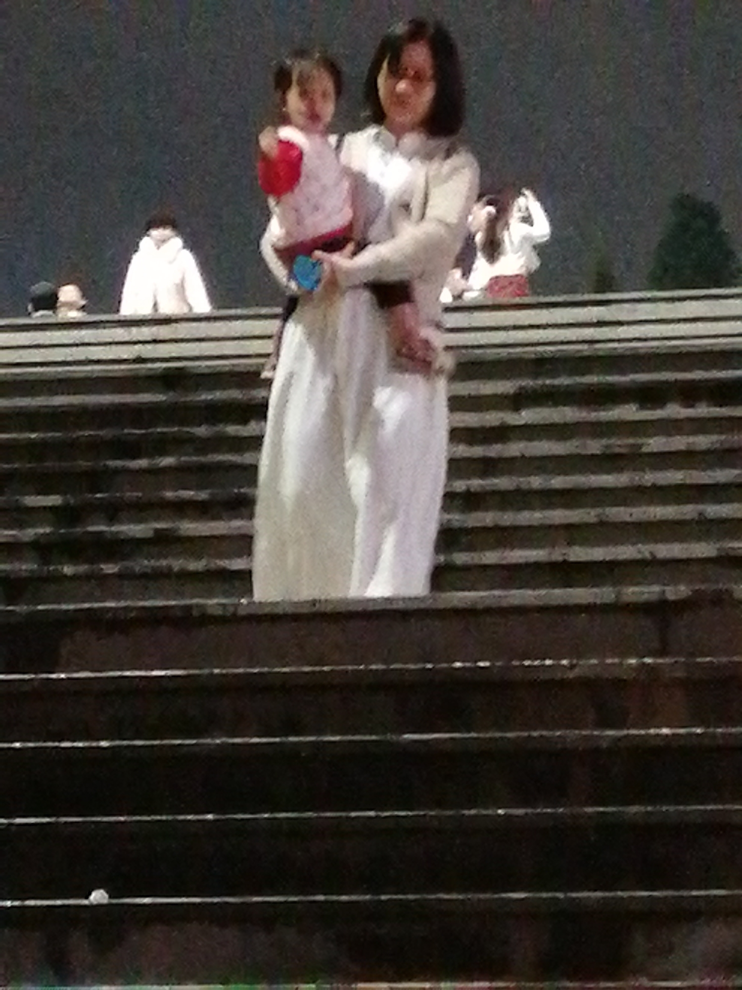 The image depicts a woman holding a young child while standing on a set of wide, dark steps. The woman, with dark hair and glasses, is wearing a light-colored top and a long, flowing white skirt. She appears to be looking forward, and her posture suggests a sense of calm. The child, held in her arms, is wearing a pink and white garment. The background is blurred, but suggests an outdoor setting at dusk or nighttime, with some indistinct figures in the distance, one of whom is wearing a white coat. The steps are made of a dark material and appear to be outdoors, possibly in a public space like a park or monument. The overall lighting is dim, indicating that the photo was taken in low-light conditions. There is no visible text in the image.