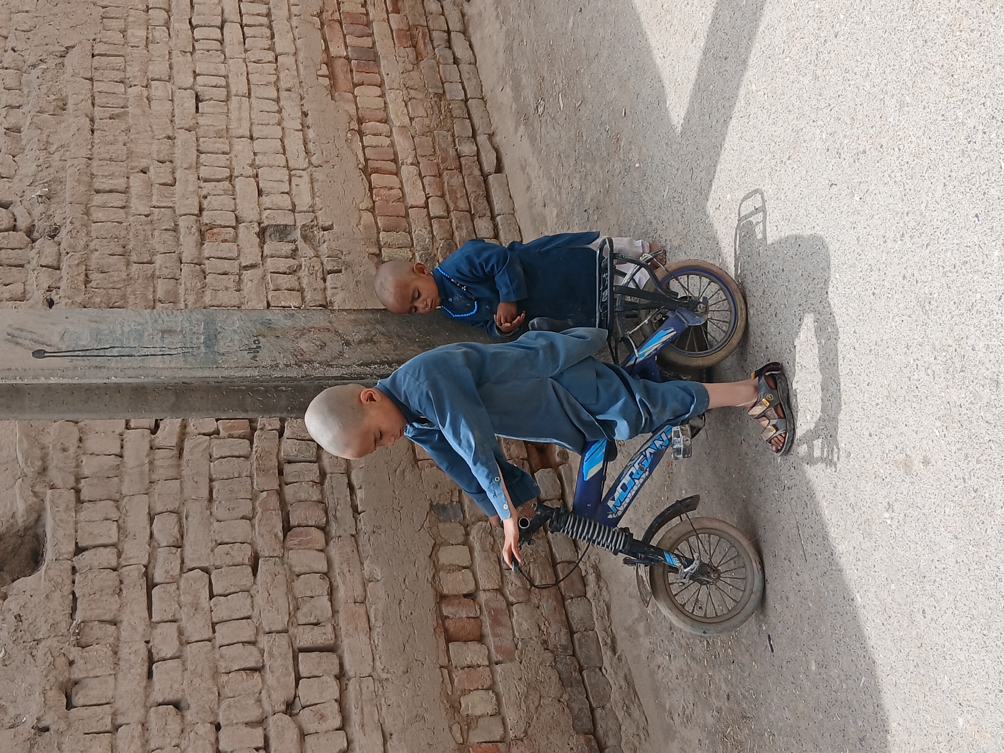 Two young boys in blue traditional clothing are next to a brick wall on a concrete surface. One boy is lying down next to the wall, while the other is near a bicycle. The scene is located in Quetta City Tehsil, Pakistan.