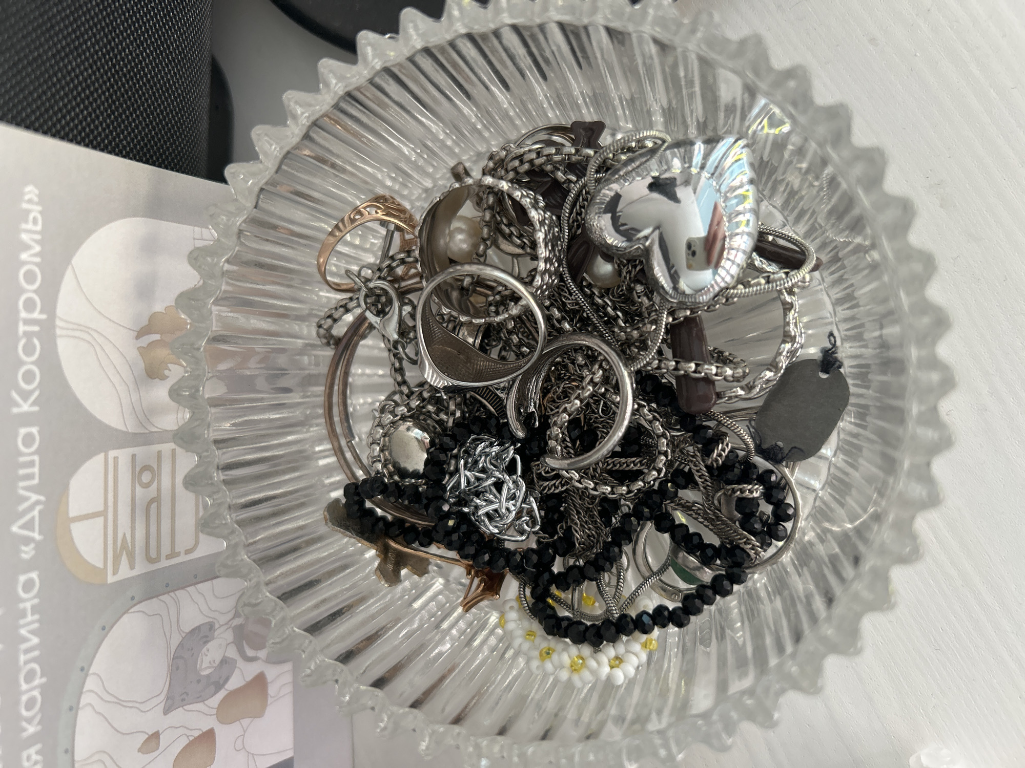 This is a top-down, close-up shot of a clear, fluted glass dish filled with an assortment of jewelry. The dish is full of various silver-toned necklaces, bracelets, and rings, some of which are adorned with pearls or black beads. A prominent heart-shaped pendant with a reflective surface is visible near the center of the dish, showing a blurred reflection of what appears to be a hand holding a phone. Some of the chains are tangled together, creating a dense collection of metallic elements. Among the silver pieces, there are a few gold-toned rings and a dark gray, dog-tag-like pendant. A small, delicate bracelet made of white and yellow beads, resembling small flowers, is also present.
To the left of the dish, part of a black textured object, possibly a speaker or a piece of electronics, is visible. Next to it is a portion of a light-colored surface with text in a Cyrillic alphabet. The visible text reads "я картина «Душа Костромы»". The lighting suggests it is daytime, with natural light illuminating the scene from the right, casting shadows within the dish and on the white surface it rests upon. The overall impression is one of collected personal adornments, casually displayed.