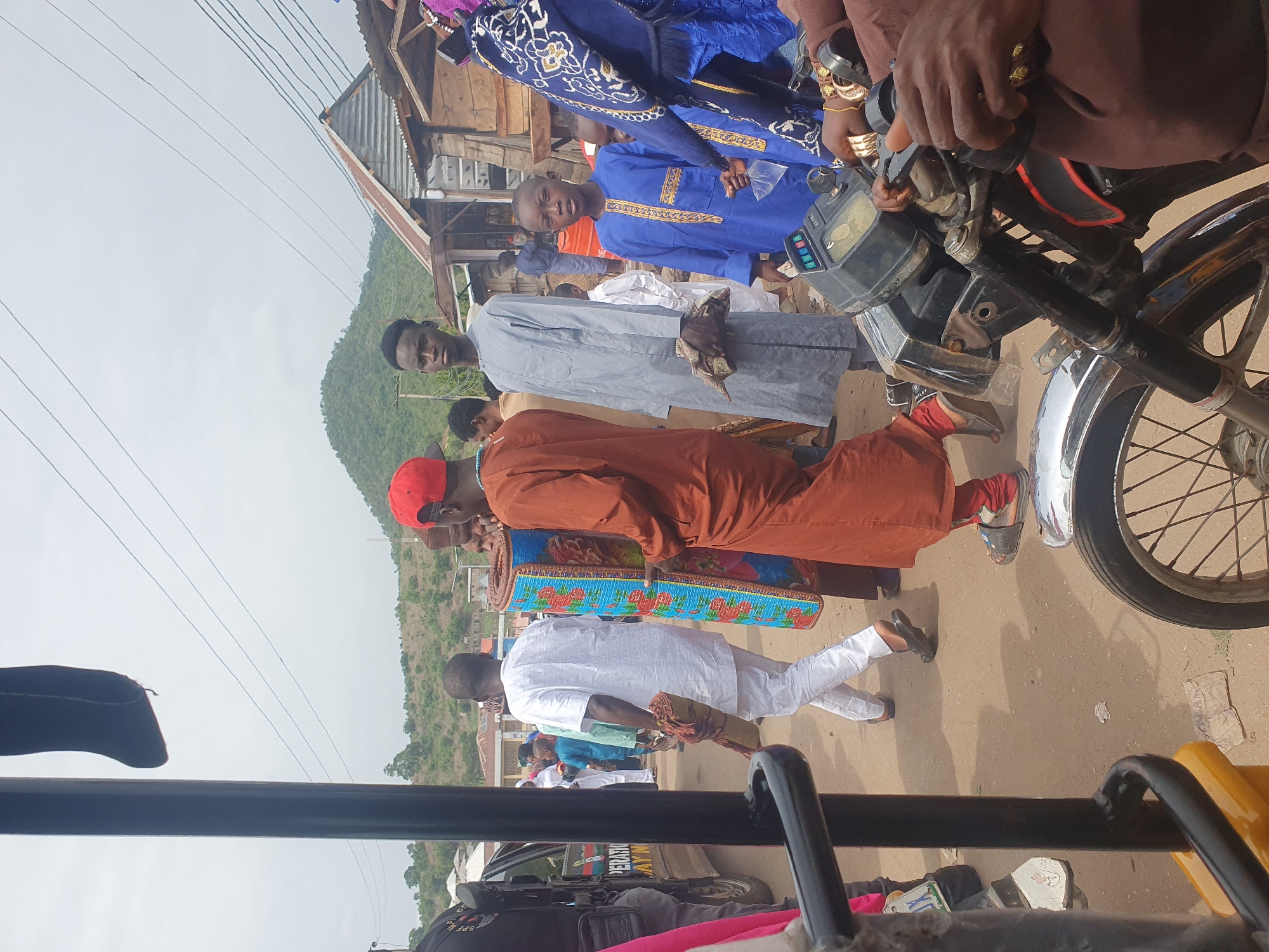 A group of people are walking along a paved road. A motorcycle is parked on the right side of the road. The people are dressed in various clothing, including traditional attire. One person is wearing a red cap and carrying a rolled-up item with a blue and red design. In the background, there is a hill and a building with a corrugated metal roof. The image appears to be taken from inside a vehicle, possibly a taxi or a similar type of public transport. Part of the vehicle's interior is visible in the foreground. The location is likely a street in Lokoja, Nigeria, given the background and clothing styles.