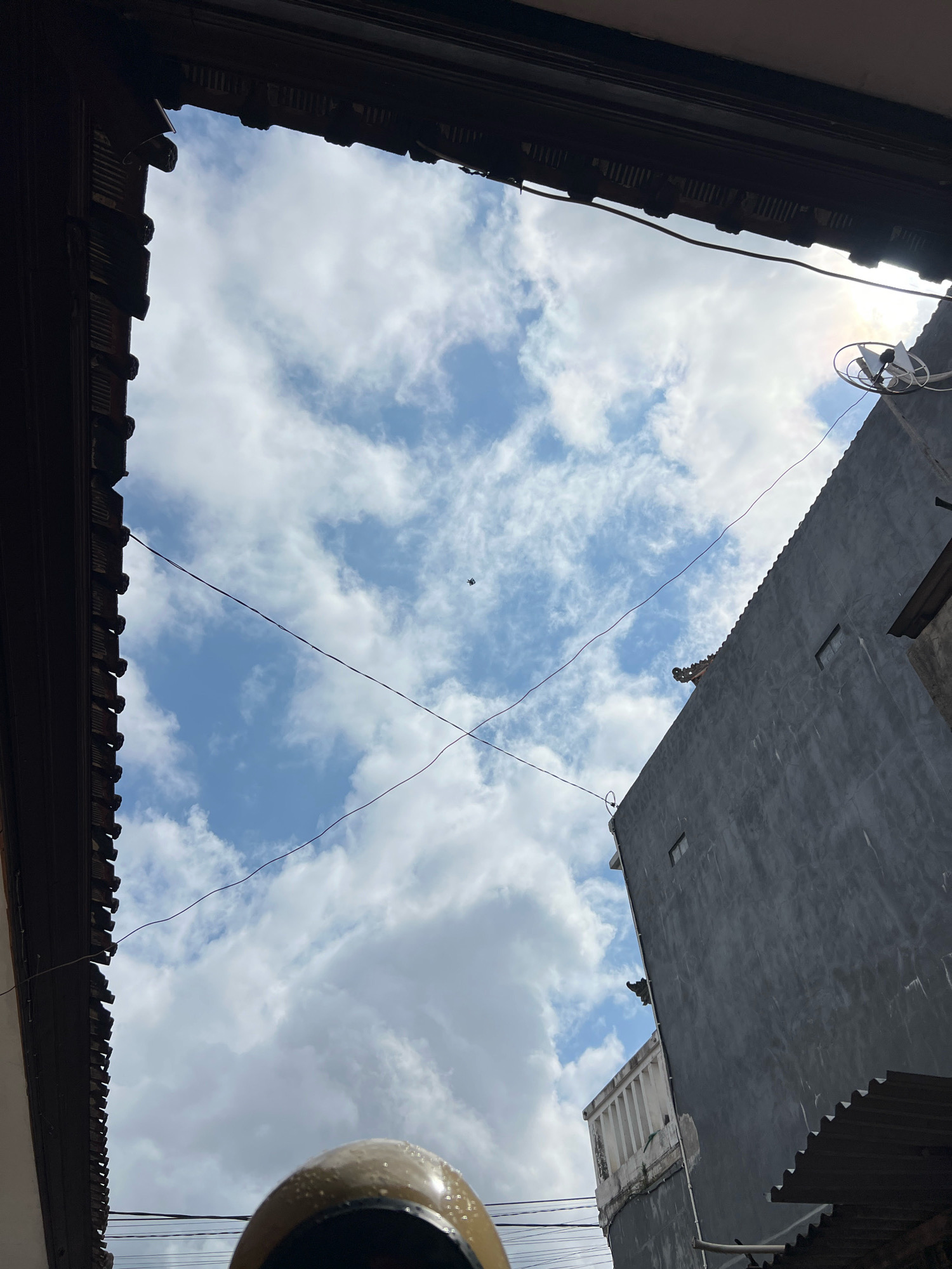 This is a slightly upward-angled photograph of a sky visible through an opening framed by buildings and a roof overhang. The sky is blue with scattered white clouds. In the upper right portion of the image, a satellite dish is mounted on the side of a grey, multi-story building. Wires are strung across the sky. A small, dark object, possibly a bird or insect, is visible in the sky to the right of the center. In the bottom foreground, the top of a light-colored helmet is visible. The scene appears to be in an urban environment, likely Denpasar, Indonesia, given the architectural style of the buildings and the presence of outdoor wiring.