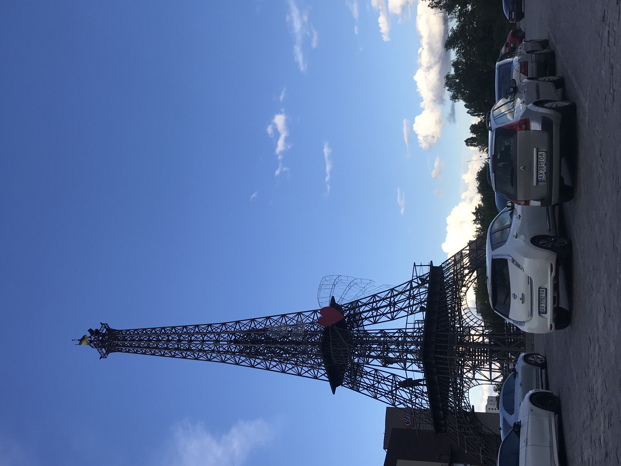 The image captures a replica of the Eiffel Tower in Kharkiv, Ukraine. The tower is a metal lattice structure against a clear blue sky with scattered clouds. A red heart is affixed to the tower. Parked cars line the adjacent street. A partial view of a building with the word "City" is visible. The Ukrainian flag flies atop the tower.