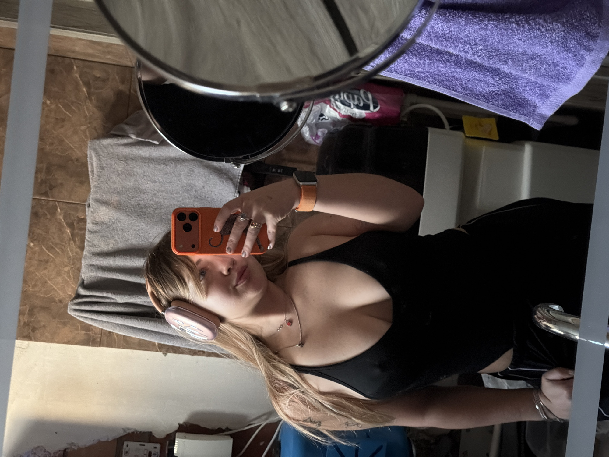 An indoor mirror selfie shows a young woman with long blonde hair, looking directly into the camera of her orange smartphone, which she holds up to her face. She is wearing a black tank top and what appear to be black shorts or pants. Pink headphones rest on her head. Her left wrist has an orange Apple Watch, and she wears multiple rings on her left hand. A necklace with a heart pendant and another charm is visible around her neck.
The reflection reveals a tiled floor in shades of brown and beige. Various household items are scattered in the background: a grey towel on the floor, a purple towel hanging or folded, a black round object (possibly a speaker), a white sink or basin, and a blue container. A pink package with the brand "Babyliss" is discernible behind the black object. Near the white basin, a small yellow item shows what looks like the number "17". The mirror itself has a visible chrome handle or frame on the right side. The overall lighting suggests an indoor setting, possibly a bathroom, with no specific time of day indicated.