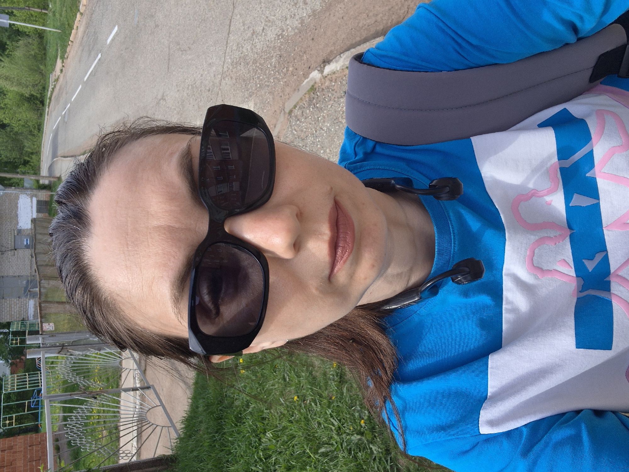 A woman wearing sunglasses, a blue and white shirt, and a gray backpack strap is standing outside in an urban area. The background includes a road, a building, a metal fence, and a grassy area with yellow flowers. The location is in the city of Unknown, Unknown.