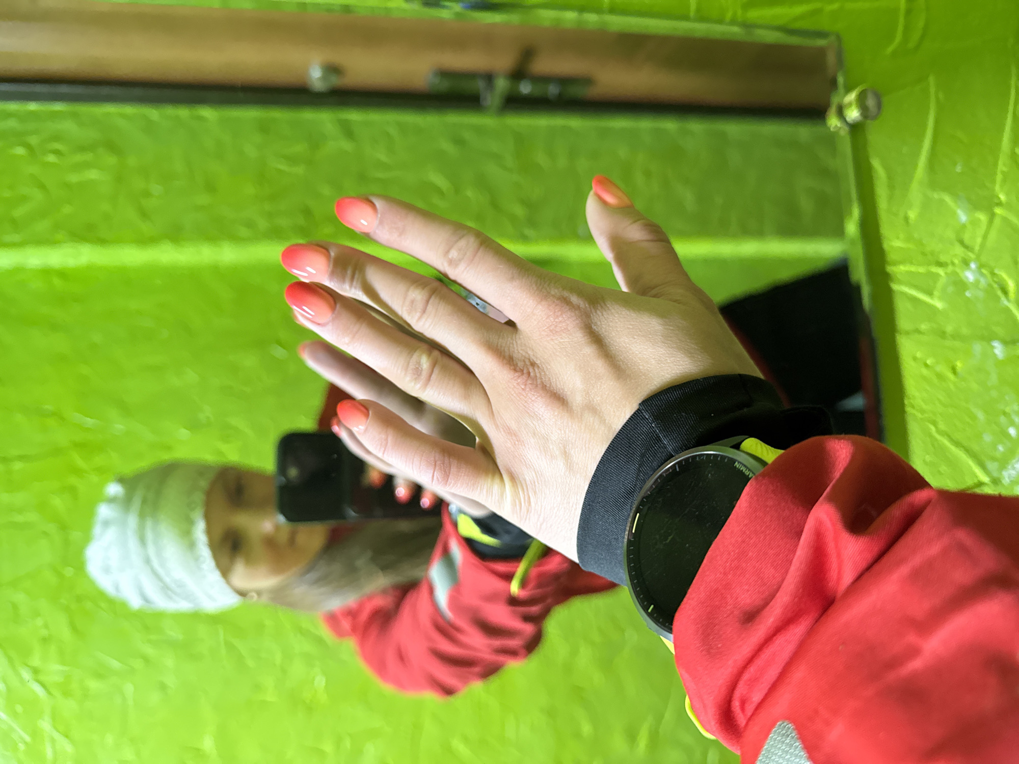 A selfie of a person wearing a red jacket, white beanie and Garmin smartwatch is reflected in a mirror. The person's hand with an ombre manicure is in the foreground. The background is a green wall. The location is Chaikovsky, Russia.