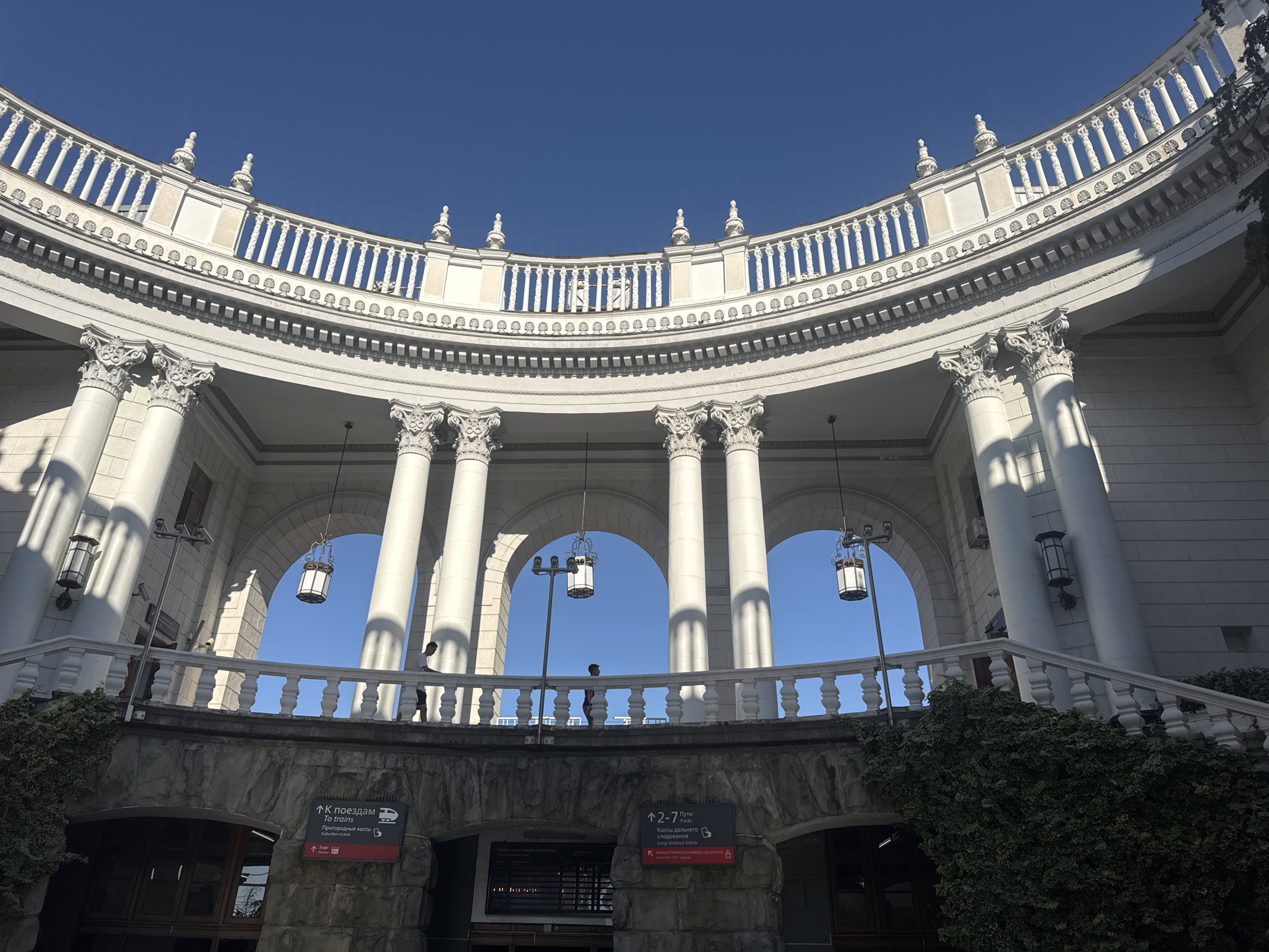 This image captures a wide-angle view of the facade of a grand, semicircular building, likely a train station, in Sochi, Russia. The architecture is classical, featuring a series of large white columns supporting an ornate, curved balustrade. Above this, a decorative railing with finials lines the top of the structure. Several lanterns hang from the arches, and in the distance, the clear blue sky indicates a bright, sunny day.
The scene is set in the daytime, with strong sunlight casting distinct shadows from the columns and architectural elements onto the building's surfaces. Two people are visible on the upper walkway, one in silhouette and another further back, suggesting a leisurely or transitional moment. Below the main structure, a stone-faced lower level is visible with entrances.
Several signs with text in both Russian and English provide directional information. One sign indicates "↑ К поездам / To trains" and "Пригородные кассы / Suburban tickets". Another sign points to "↑ 2-7 Пути / Tracks 2-7" and "Кассы дальнего следования / Long-distance tickets". There is also a sign for "automatic luggage storage" or "automatic storage lockers". The presence of these signs and the architectural style strongly suggest this is a transportation hub, specifically a train station. The overall atmosphere appears calm and orderly, characteristic of a functional public space on a pleasant day. Lush green ivy climbs the lower sections of the building, adding a touch of nature to the stone and classical architecture.
