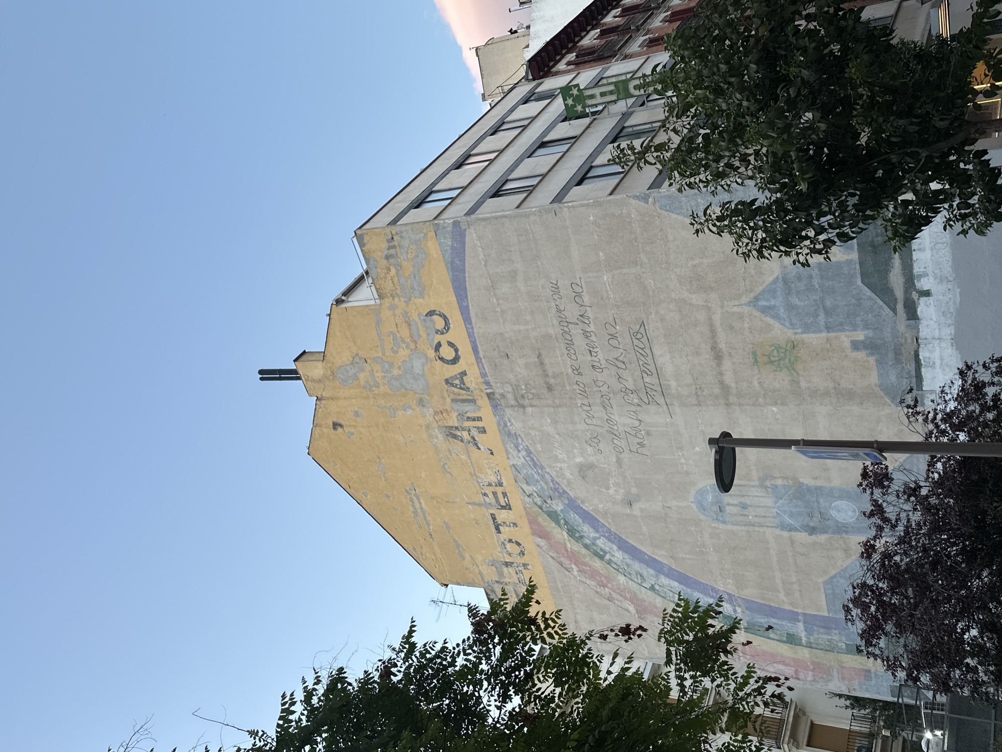 The image depicts the side of a building in Madrid, Spain. Painted on the building is a faded advertisement for the "Hotel Anaco." A curved blue and green band, possibly part of a larger design, surrounds the hotel name. The rest of the wall surface is covered in inscriptions in Spanish. A streetlight obscures the lower right part of the painting. Trees occupy the foreground, framing the building and obscuring the view. The sky above is a clear light blue.