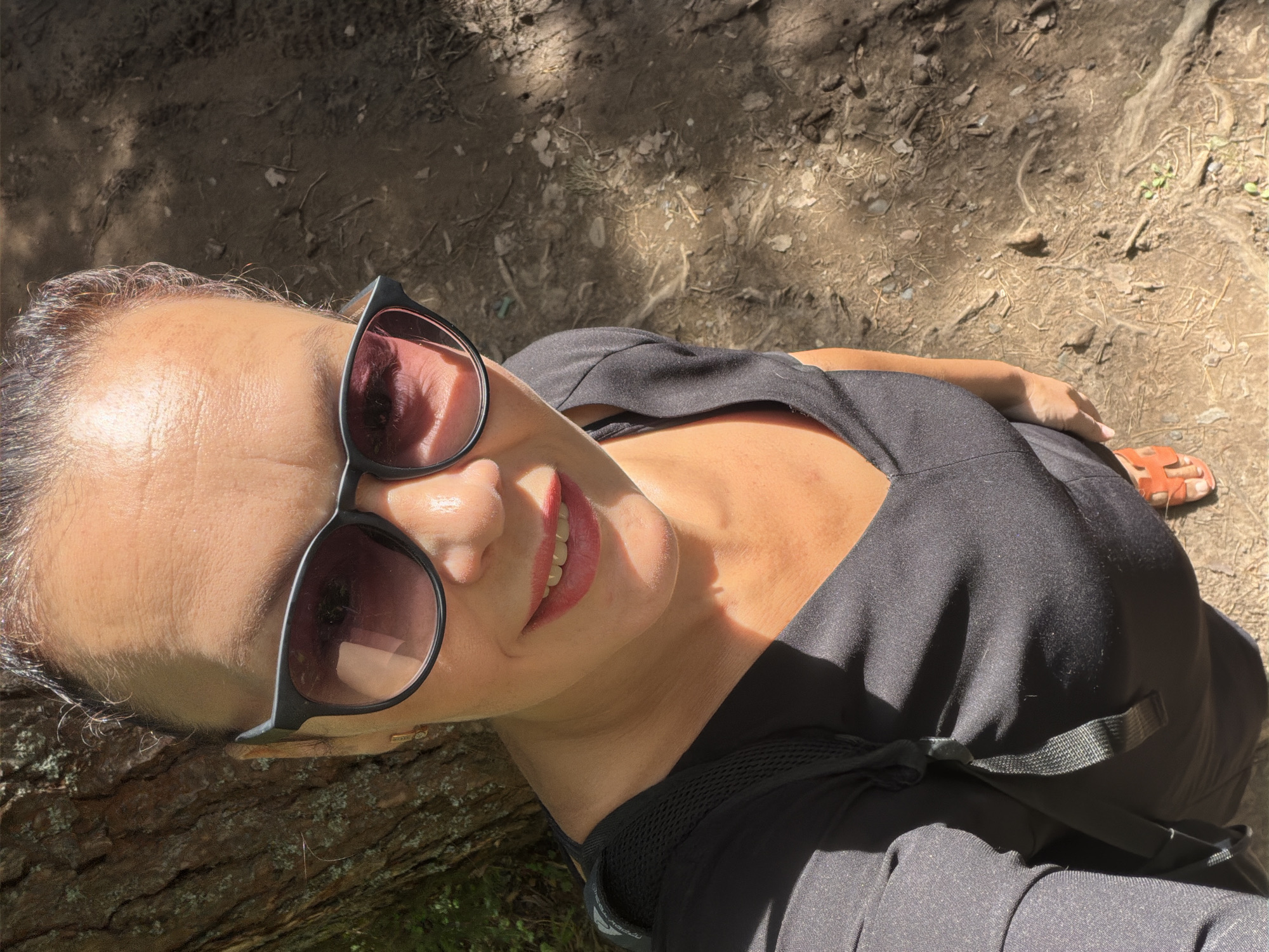 A woman is taking a selfie outdoors, leaning against a tree. She is wearing a black V-neck top and sunglasses. The background shows a dirt path with some dry leaves and small plants. The lighting suggests it is a sunny day. There is no discernible information in the image that places it in Votkinsk, Russia.