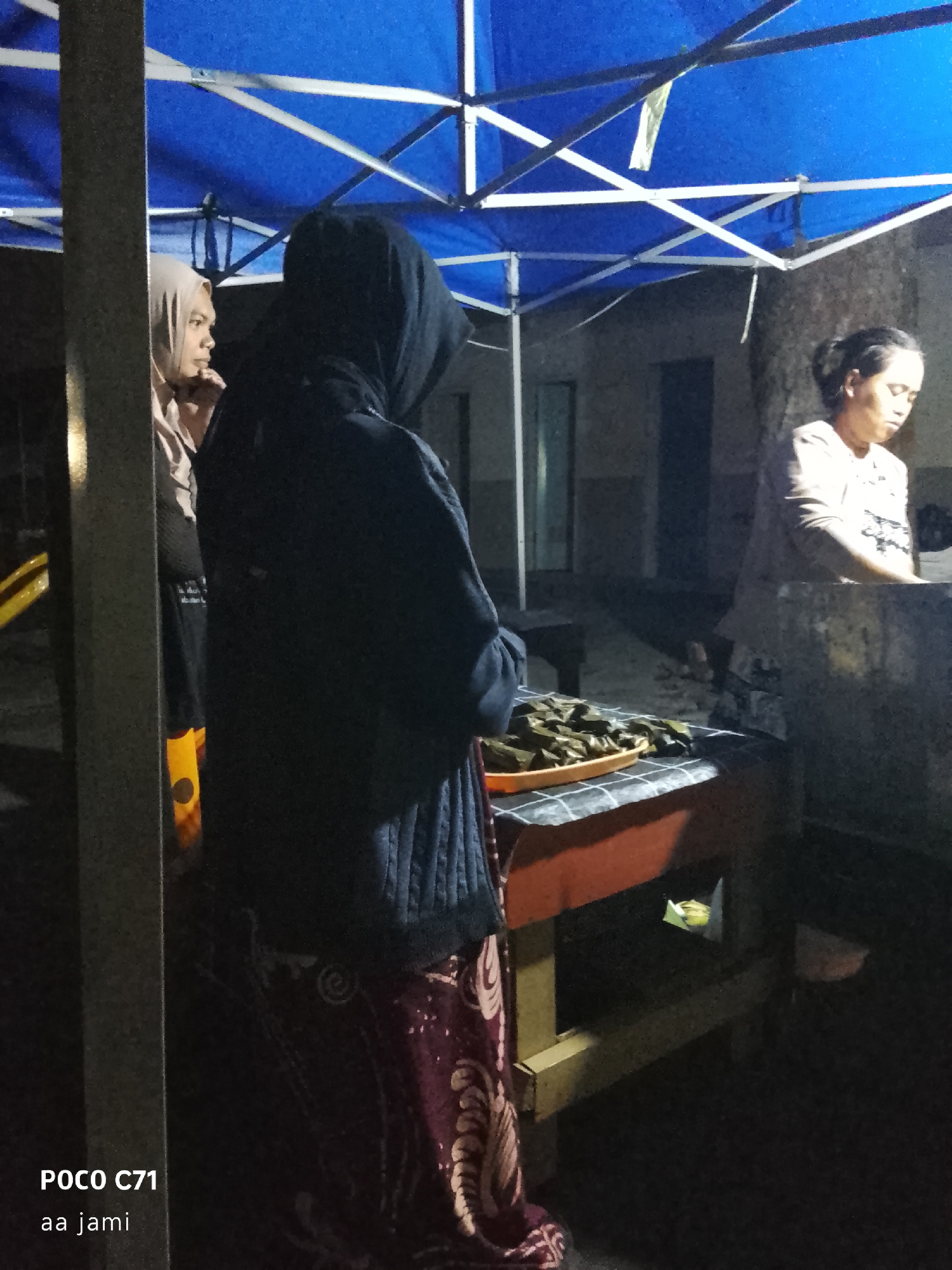 The image depicts an outdoor food stall at night, likely in Tasikmalaya, Indonesia, under a blue canopy. In the foreground, to the left, a person wearing a light-colored hijab is partially visible, facing towards the right. To their right, another person, fully cloaked in a dark hooded garment, stands facing a table covered with a checkered cloth. On this table, an orange tray holds numerous small, green, wrapped food items. The background shows a dimly lit building and other indistinct structures. To the far right, a woman with her hair pulled back is visible, leaning over a counter, appearing to be tending to the stall. The overall lighting is dim, suggesting it is nighttime, and the scene has a common street food stall ambiance.
