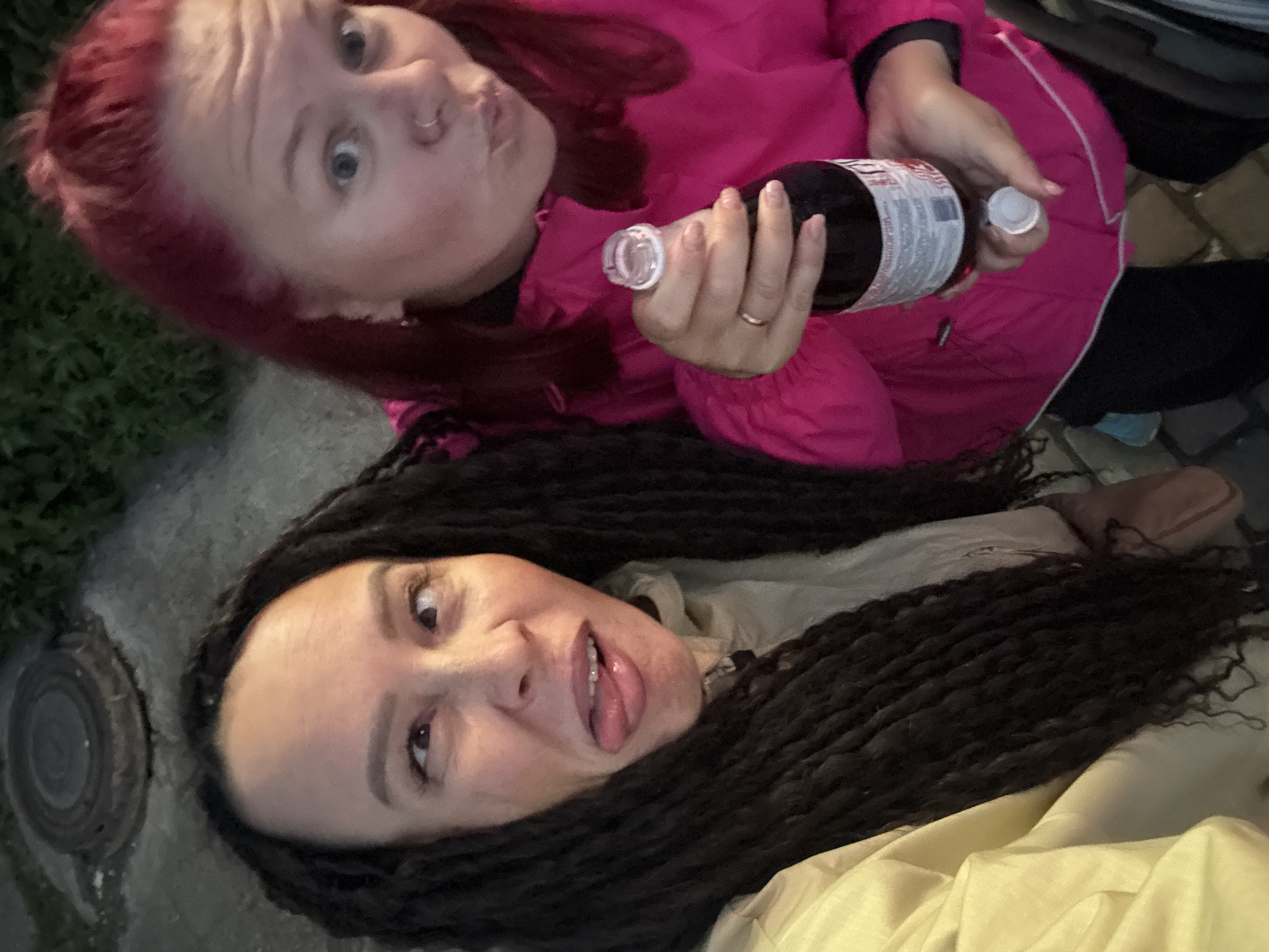 The scene takes place outdoors in the city of Chaikovsky, Russia, depicting two individuals engaged in a playful pose. On the left, a female individual with long, dark dreadlocks or braids is seen from the chest up, wearing a light-colored jacket. She is making a humorous expression with her eyes crossed and tongue extended. To her right, another female individual with bright red hair, pulled back from her face, is visible from the waist up, wearing a bright pink hooded jacket. She has a metallic ring piercing in her left nostril and a gold-colored ring on her left ring finger. This individual is holding a clear glass bottle containing a dark liquid, with its white cap removed and held in her left hand. The bottle has a white label with red text. She is looking directly at the camera with a slight pout. The background consists of dark paving stones or concrete, with a round metal manhole cover visible behind the left individual's head. Green foliage is faintly discernible in the upper background. The lighting suggests dusk or nighttime conditions.