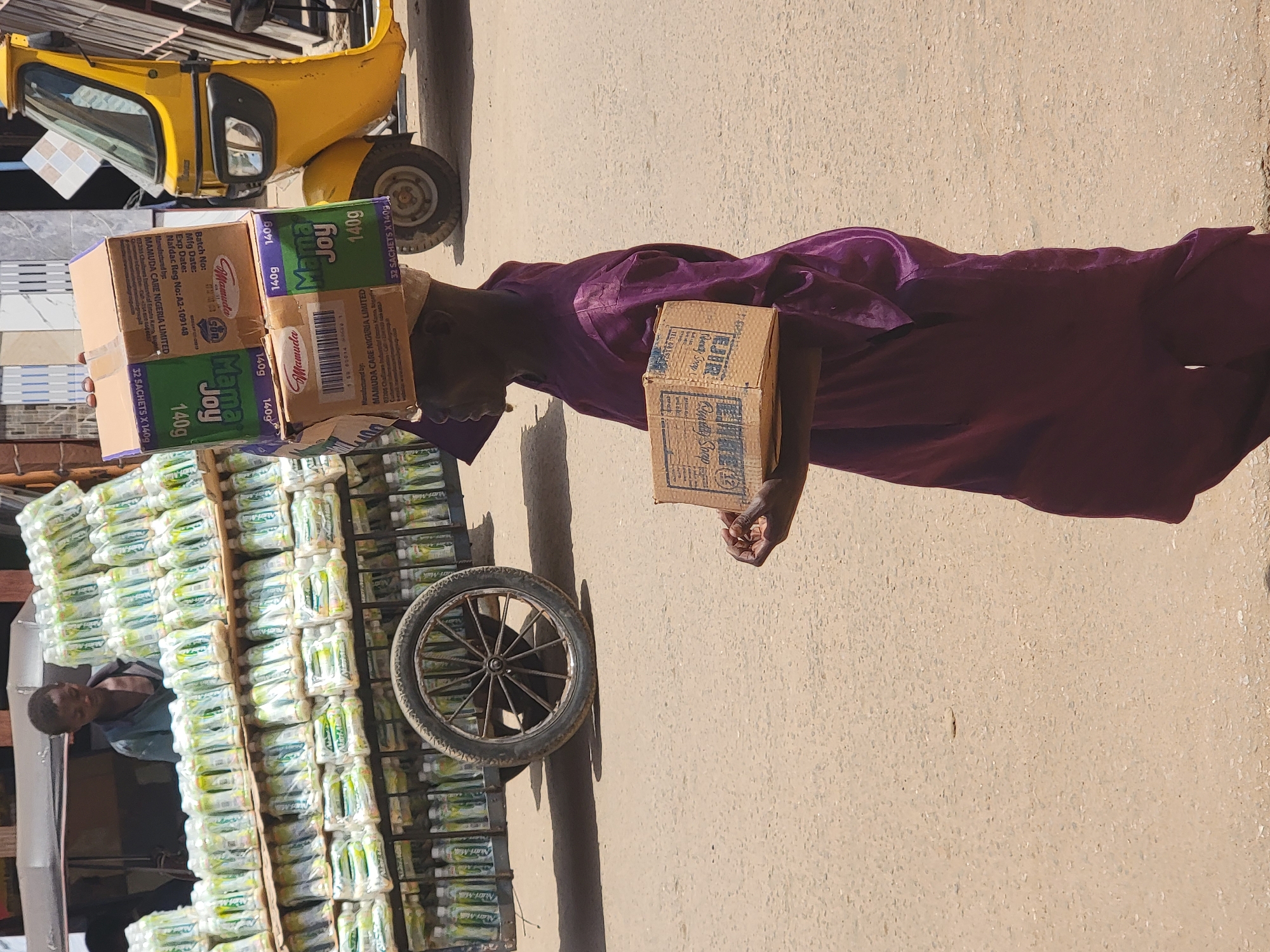 A man dressed in a plum-colored robe carries boxes of "Mama Joy" brand food supplement on his head and a box labeled "Ezir" in his right hand. He is standing next to a cart containing stacks of what appear to be the same "Mama Joy" food supplement. A three-wheeled auto rickshaw (tuk-tuk) is partially visible in the background. The scene is set on a street in Maiburiji, Nigeria.