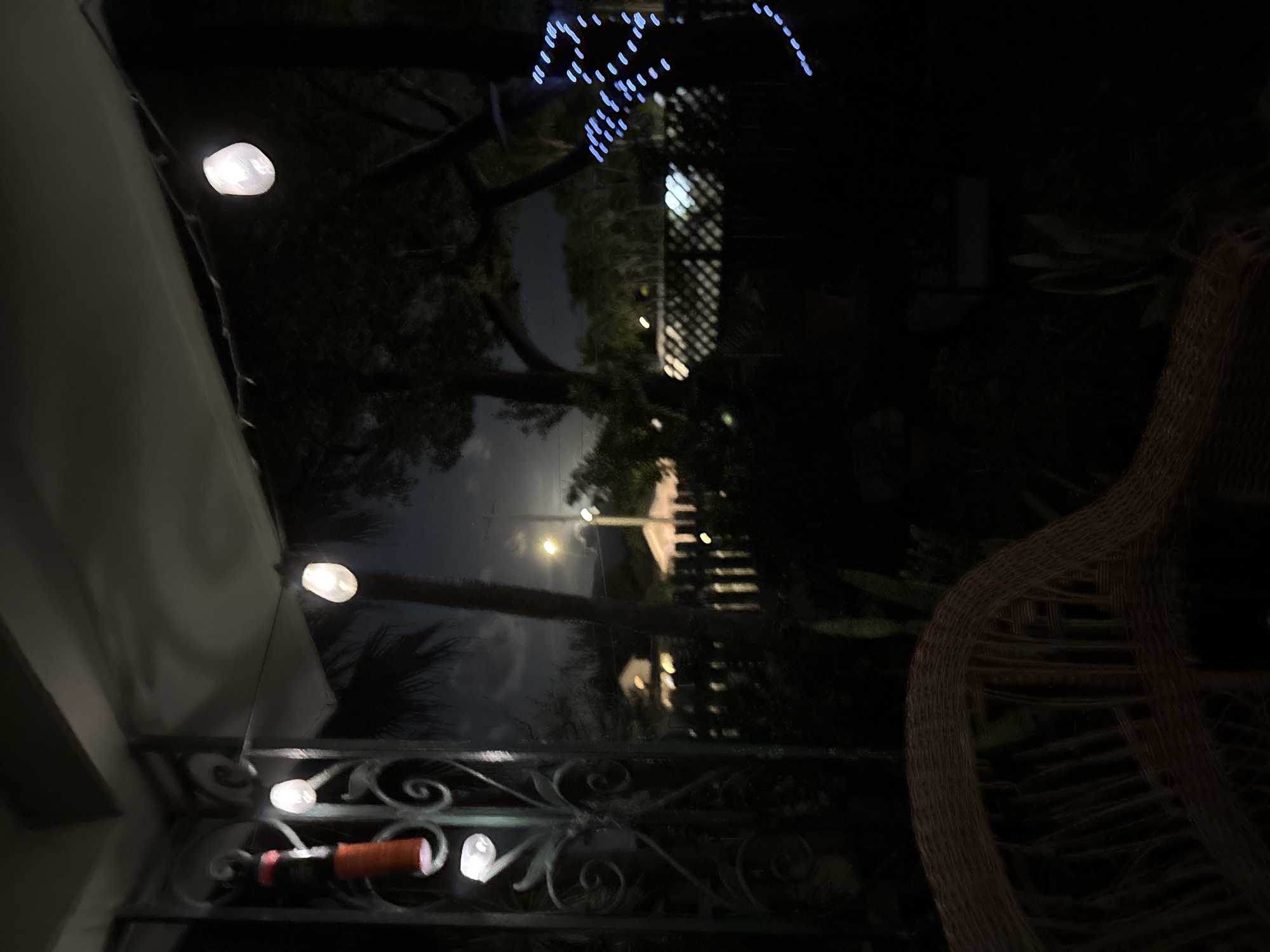 This is a nighttime photograph taken from a porch in Key West, Florida. The foreground is dominated by the armrest of a wicker chair, rendered in dark tones. Beyond the chair, a metal railing with ornate scrollwork frames the scene. A string of white globe lights extends across the porch, with several bulbs illuminated. A red object, possibly a bottle, hangs from the railing.
The background reveals a dimly lit suburban or residential area. A palm tree stands prominently in the mid-ground, silhouetted against the night sky. In the distance, houses with illuminated windows and a street light are visible. A lattice-patterned fence runs horizontally across the mid-ground. Blue string lights are visible on a tree to the right. The moon is faintly visible in the sky. The overall atmosphere is dark and tranquil.