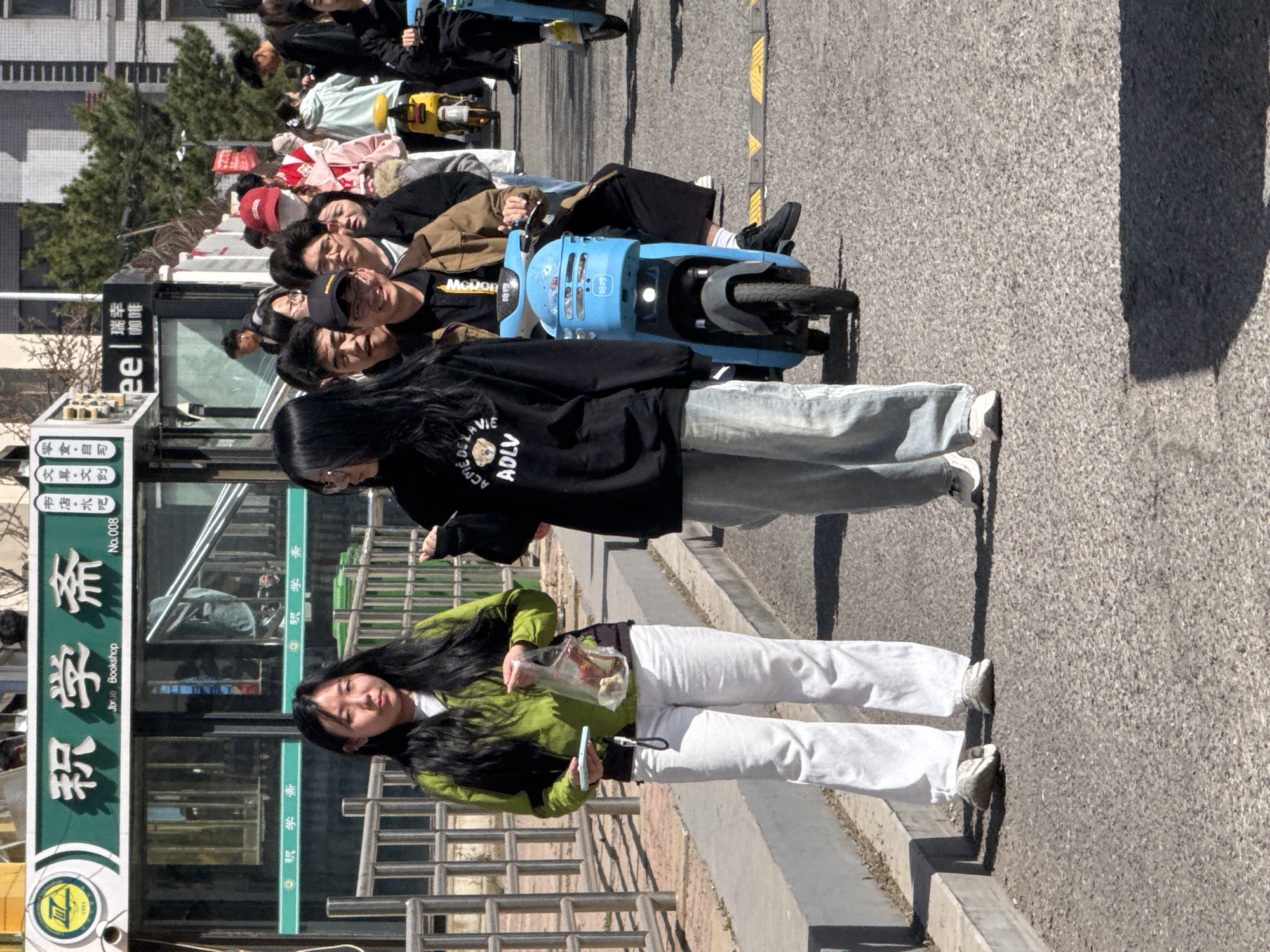 The scene captures a bustling street in Zhongtun, China. In the foreground, a young woman with long, dark hair is standing next to a bus stop. She is wearing a green jacket and white pants, and she is holding a plastic bag and a phone. Behind her, another woman is standing and wearing a black hoodie with the inscription "ACME DELA VIE ADLV" and light blue jeans. A line of people riding blue scooters is also visible. The bus stop, with signage in both Chinese and English, identifies the location as "Jixue Bookshop, No.008." A sign for "luckin coffee" is also visible at the bus stop. The street appears to be well-maintained and populated with trees and modern buildings.
