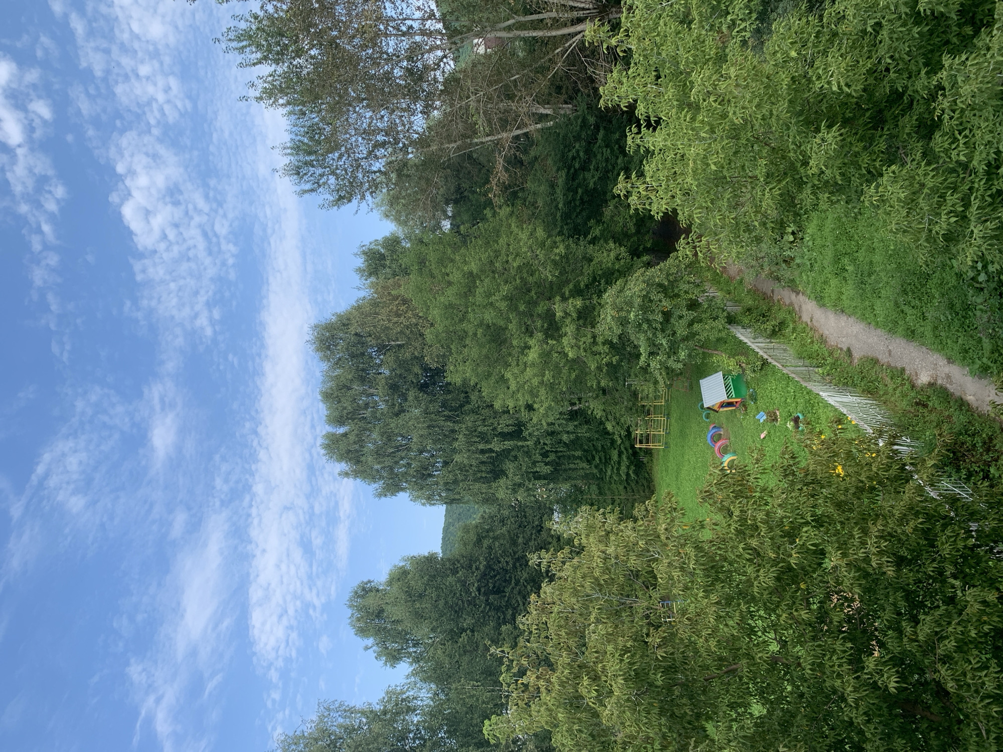 This is an aerial outdoor photograph taken on a sunny day with a bright blue sky filled with scattered white clouds, showcasing a lush green landscape. In the foreground, dense green foliage from trees dominates the left and right sides of the frame, with some branches extending into the center.
A small, fenced-in area is visible in the mid-ground, featuring a bright green playhouse with a silver roof and a yellow frame structure, possibly a swing set or climbing frame. Colorful tire swings in red, yellow, blue, and pink are arranged on the grass. Several small plants or flowers are scattered throughout the grassy area. A light-colored dirt path, bordered by grass and greenery, winds its way from the lower right corner of the image towards the fenced play area. A white picket fence separates the path and surrounding greenery from the playground.
In the background, a dense forest of various trees covers rolling hills. The trees exhibit a range of green hues, suggesting different species or varying levels of sunlight exposure. The overall impression is a serene, natural environment. The scene appears to be located in or near the city of Asha, Russia, given its typical natural landscape of forested hills and modest residential or recreational areas. No people are visible in the image.
