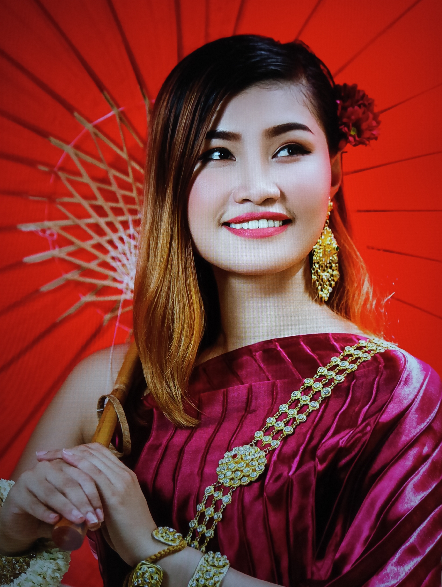 A studio portrait shows a woman holding a closed red parasol. She is adorned with gold jewelry and wearing a maroon velvet gown with gold embellishments. A red flower is tucked behind her right ear. The photograph suggests cultural or traditional attire, potentially associated with a formal event or celebration. The location is not indicated by the image. The city of Nha Trang Ward, Vietnam is not indicated in the image.