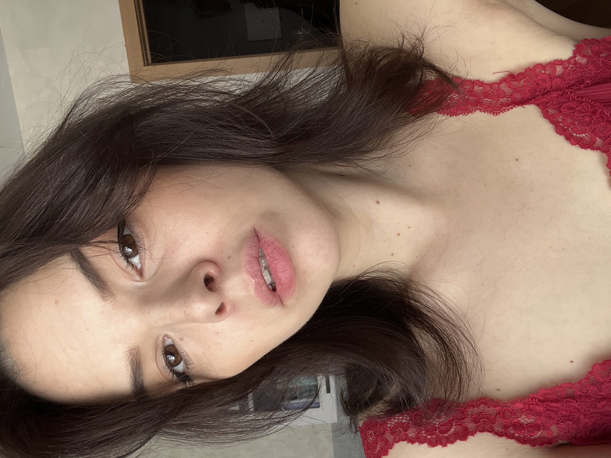 A close-up, tilted, and rotated photograph shows a woman in a red, lace-trimmed garment. The woman has long, dark brown hair and brown eyes. She is partially visible from her chest to the top of her head. Her skin has various freckles and blemishes. The background includes a wall and a window with a dark rectangular object in the frame, and possibly a partially visible room with open doorway. There is no explicit evidence to confirm a location within the city of Shushary, Russia.
