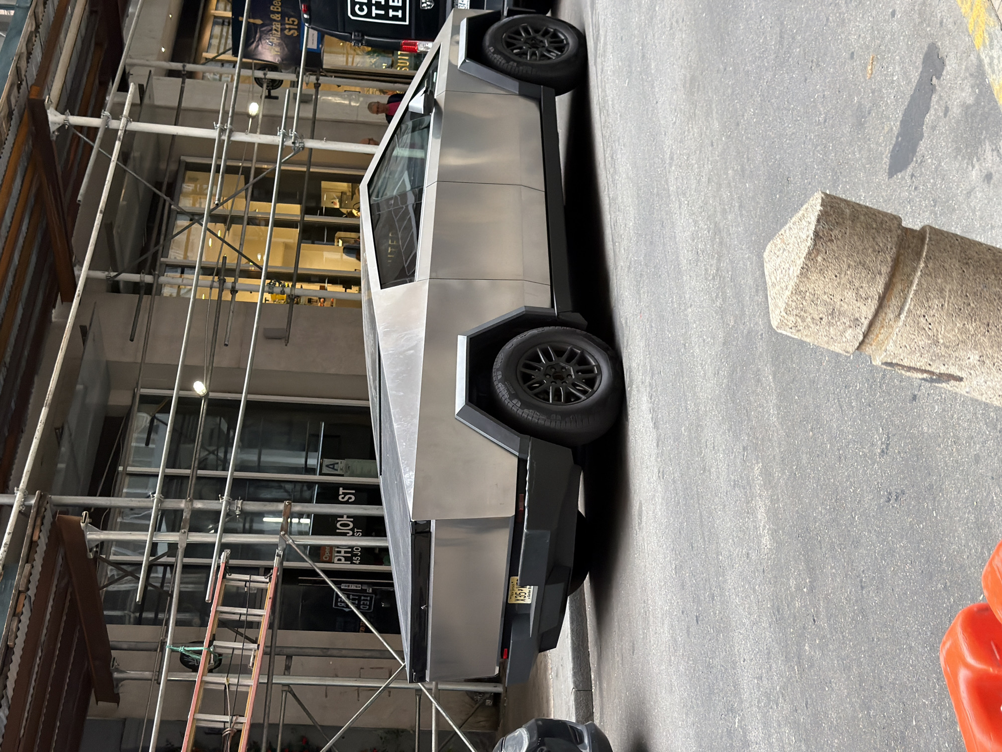 A silver Tesla Cybertruck is parked on a city street in New York, USA, next to a black van and scaffolding. The Cybertruck is facing right, and its angular, stainless steel body is visible. The license plate is visible and reads "X357M". In the background, scaffolding is erected in front of a building with large windows. A "Pizza & Beer" sign is also visible on the building. The street is paved with asphalt. A concrete bollard is in the foreground, casting a shadow. Two red plastic objects are visible in the bottom left corner of the image.